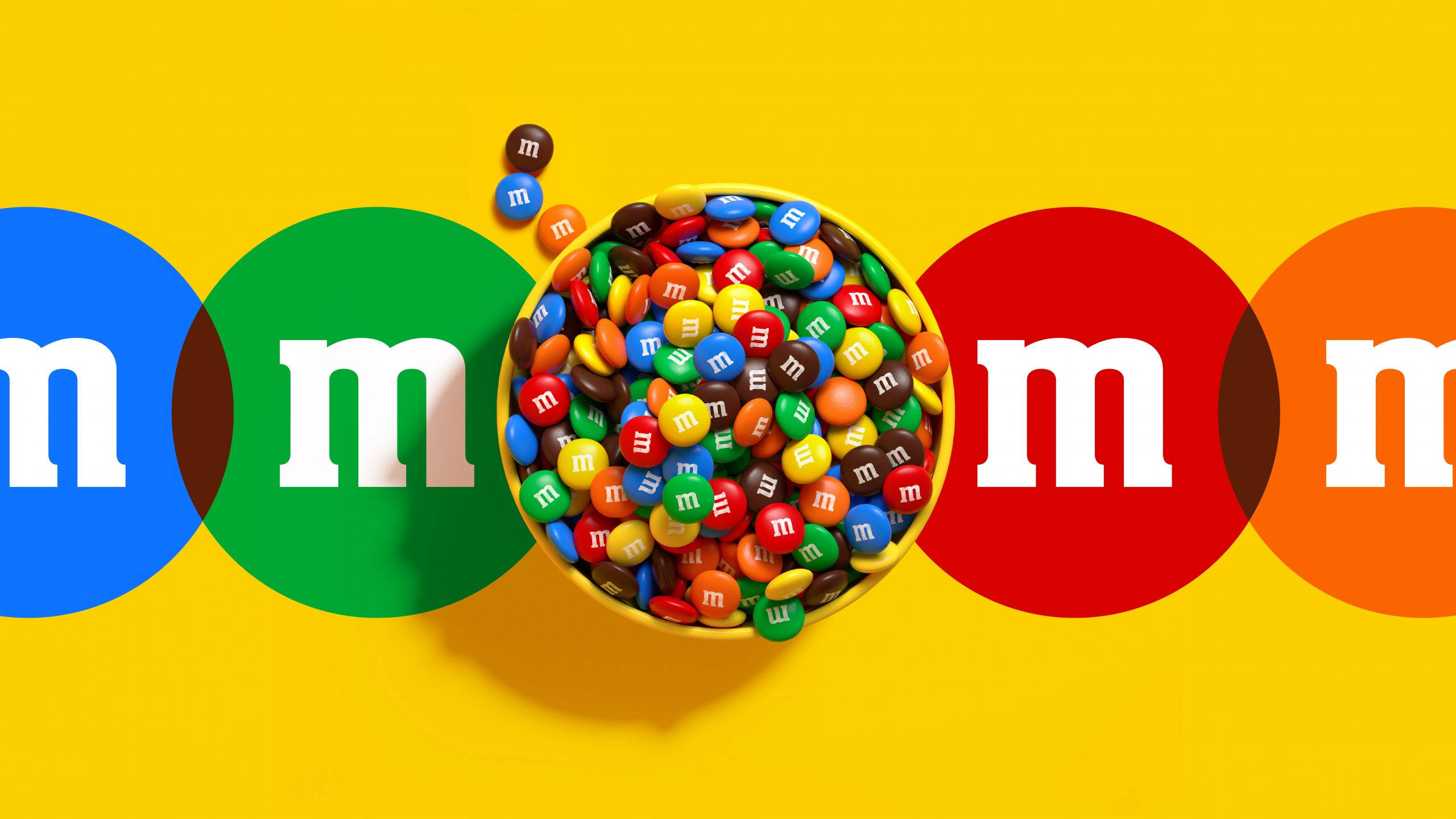 朱古力 桌布 壁紙 — 一個充滿活力的碗，裝滿了色彩繽紛的M&M's糖果，放在明亮的黃色背景上，兩側有大型品牌圓圈。