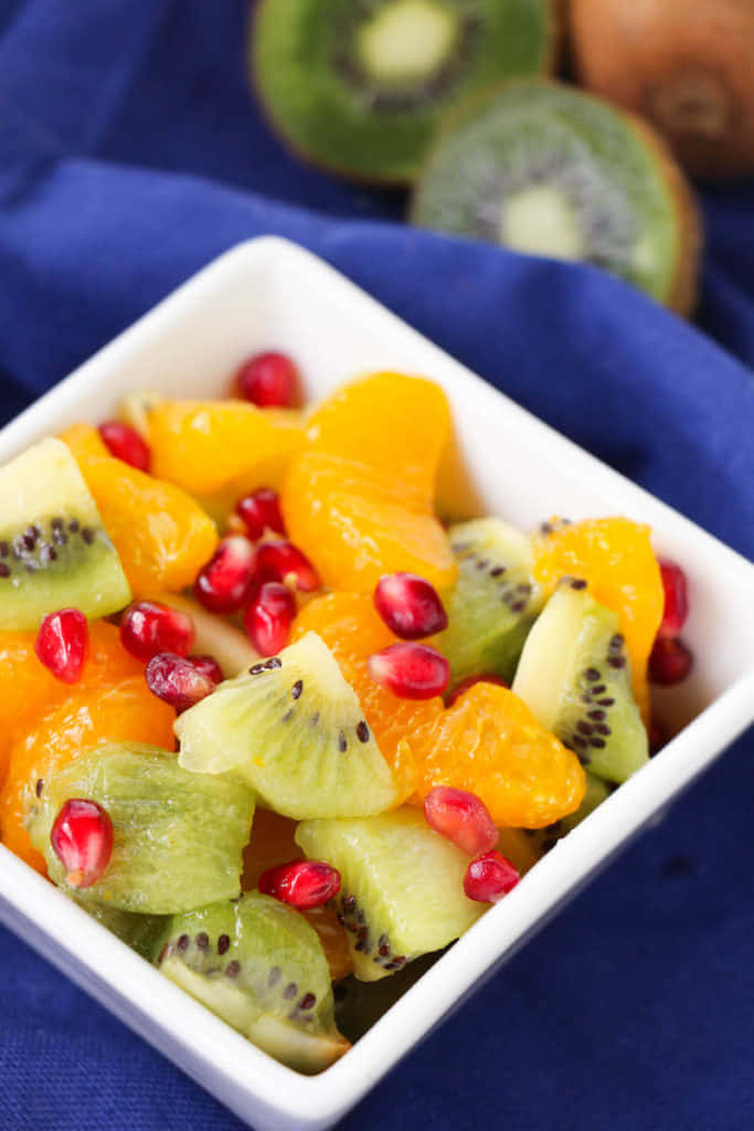 Winter Fruits 桌布 壁紙 — 一個充滿活力的碗，裡面有切片奇異果、橙瓣和石榴籽，放在深藍色布上。