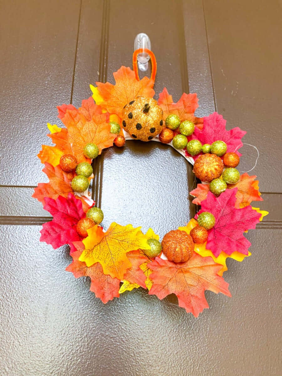 Fall Crafts 桌布 壁紙 — 一個充滿活力的秋季花環，飾有橙色、粉紅色和黃色的楓葉、閃閃發光的南瓜和綠色珠子，掛在棕色門上。