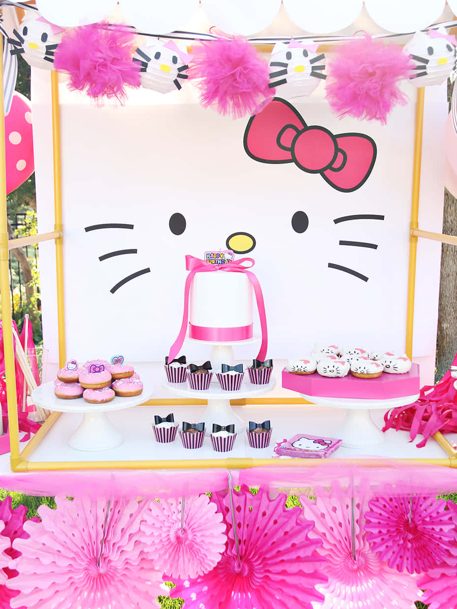 Hello Kitty Party 桌布 壁紙 — 一個充滿活力的粉紅色Hello Kitty甜品桌，上面有紙杯蛋糕、冬甩和一個蛋糕，背景是一個巨大的卡通面孔。