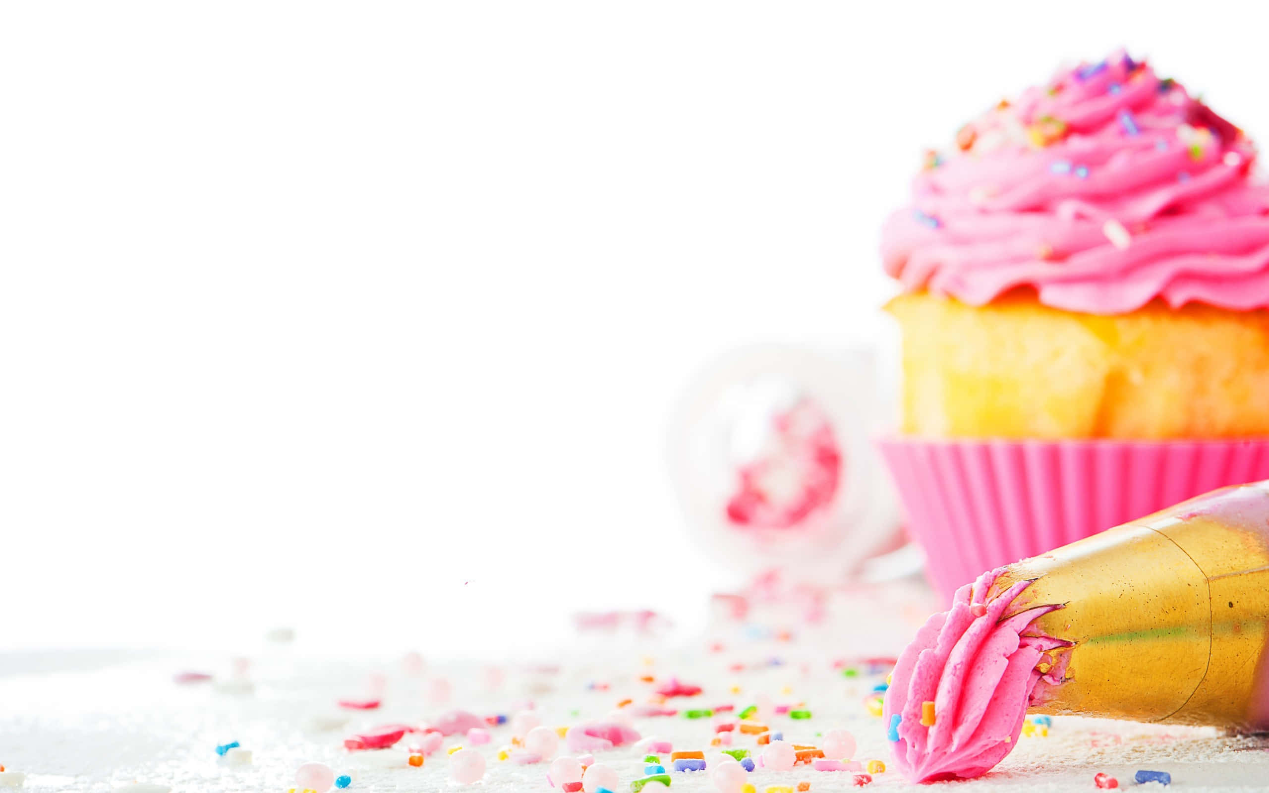 Cute Cupcake 桌布 壁紙 — 一個充滿活力的粉紅色糖霜紙杯蛋糕，上面有彩色糖粒，旁邊放著一個裱花袋，背景是明亮的白色。