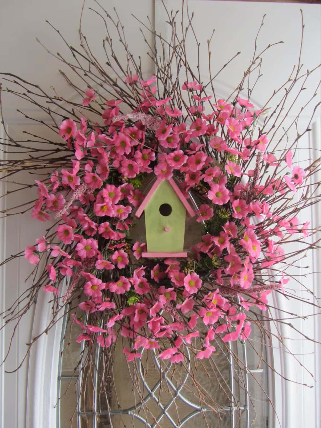 Spring Wreaths 桌布 壁紙 — 一個充滿活力的粉紅色花環，中心有一個粉綠色鳥屋，掛在帶玻璃面板的白色門上。