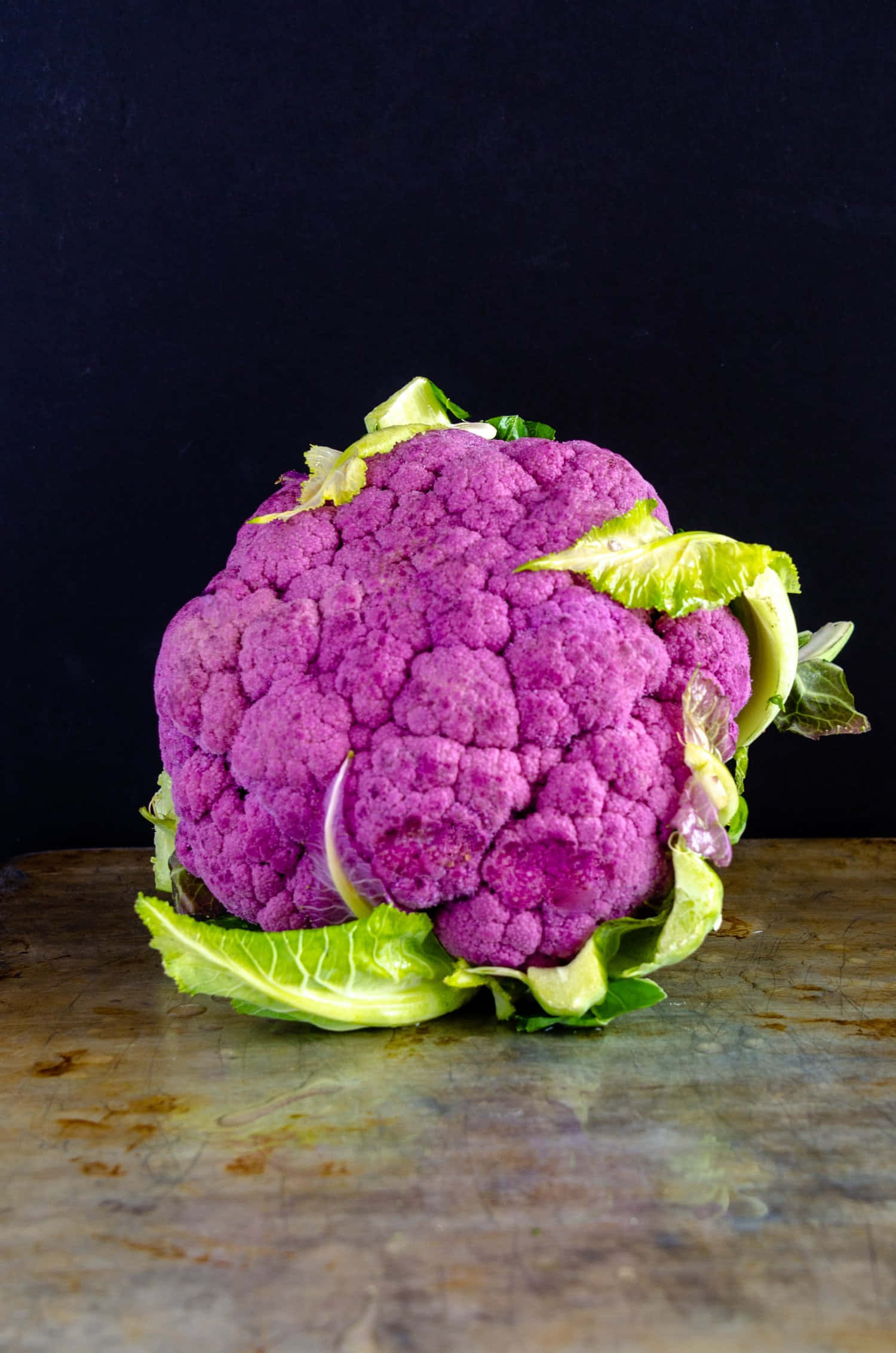 Purple Cauliflower 桌布 壁紙 — 一個充滿活力的紫色椰菜花，帶有新鮮的綠色葉子，放在一個風化的金屬表面上，背景是深色的。