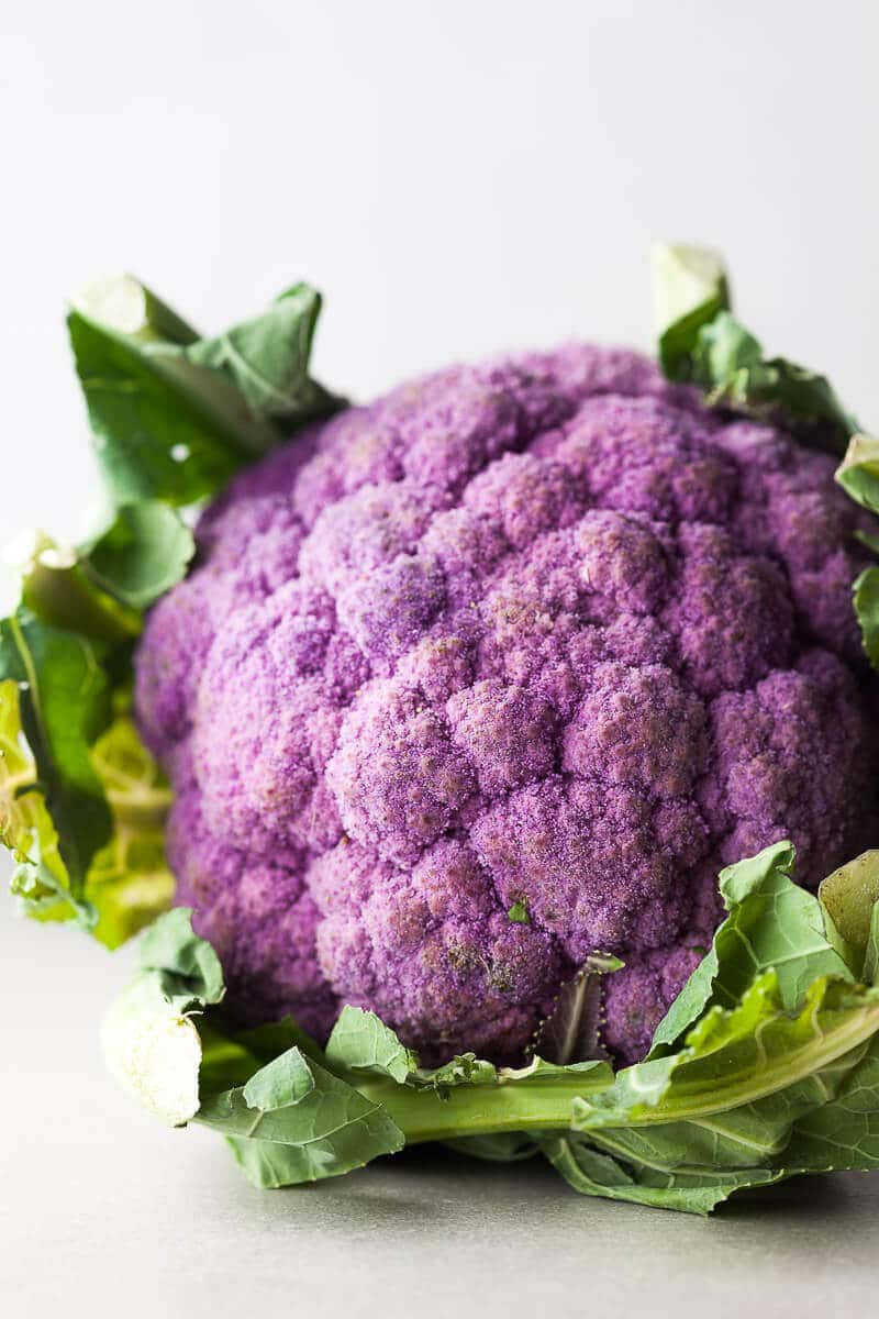 Purple Cauliflower 桌布 壁紙 — 一個充滿活力的紫色椰菜花，帶有新鮮的綠色葉子，在柔和的工作室燈光下放置在灰色中性表面上。
