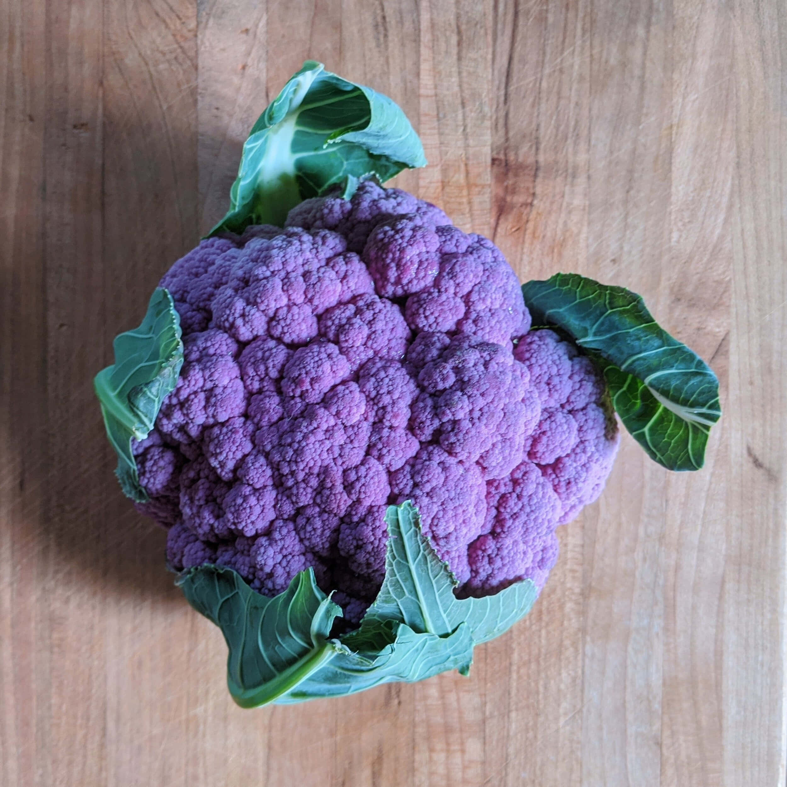 Purple Cauliflower 桌布 壁紙 — 一個充滿活力的紫色椰菜花，帶有綠色葉子，放在溫暖的木砧板上。