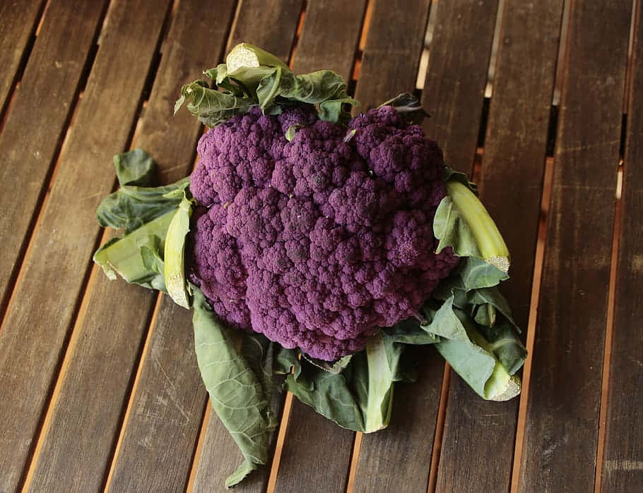 Purple Cauliflower 桌布 壁紙 — 一個充滿活力的紫色椰菜花，帶有綠色葉子，放在風化的木條表面上。