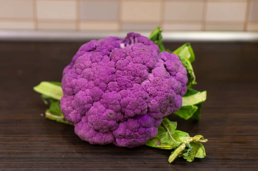 Purple Cauliflower 桌布 壁紙 — 一個充滿活力的紫色椰菜花，帶有綠色葉子，放在深色木質枱面上，背景是瓷磚牆。