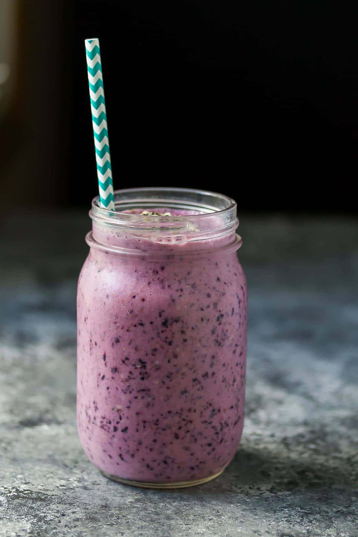 Blueberry Smoothie 桌布 壁紙 — 一個充滿活力的紫色莓果冰沙放在玻璃罐中，配上一根青綠色和白色條紋的吸管，置於有紋理的灰色表面上。