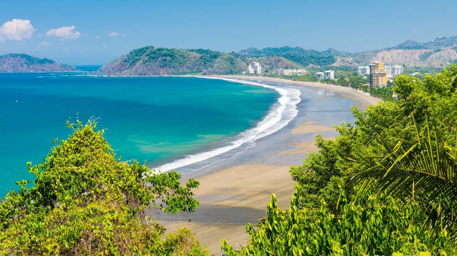 Costa Rica 桌布 壁紙 — 一個充滿活力的綠松石海灣沿著沙灘蜿蜒，兩旁是茂密的綠色山丘和現代化的沿海建築，在晴朗的藍天下。
