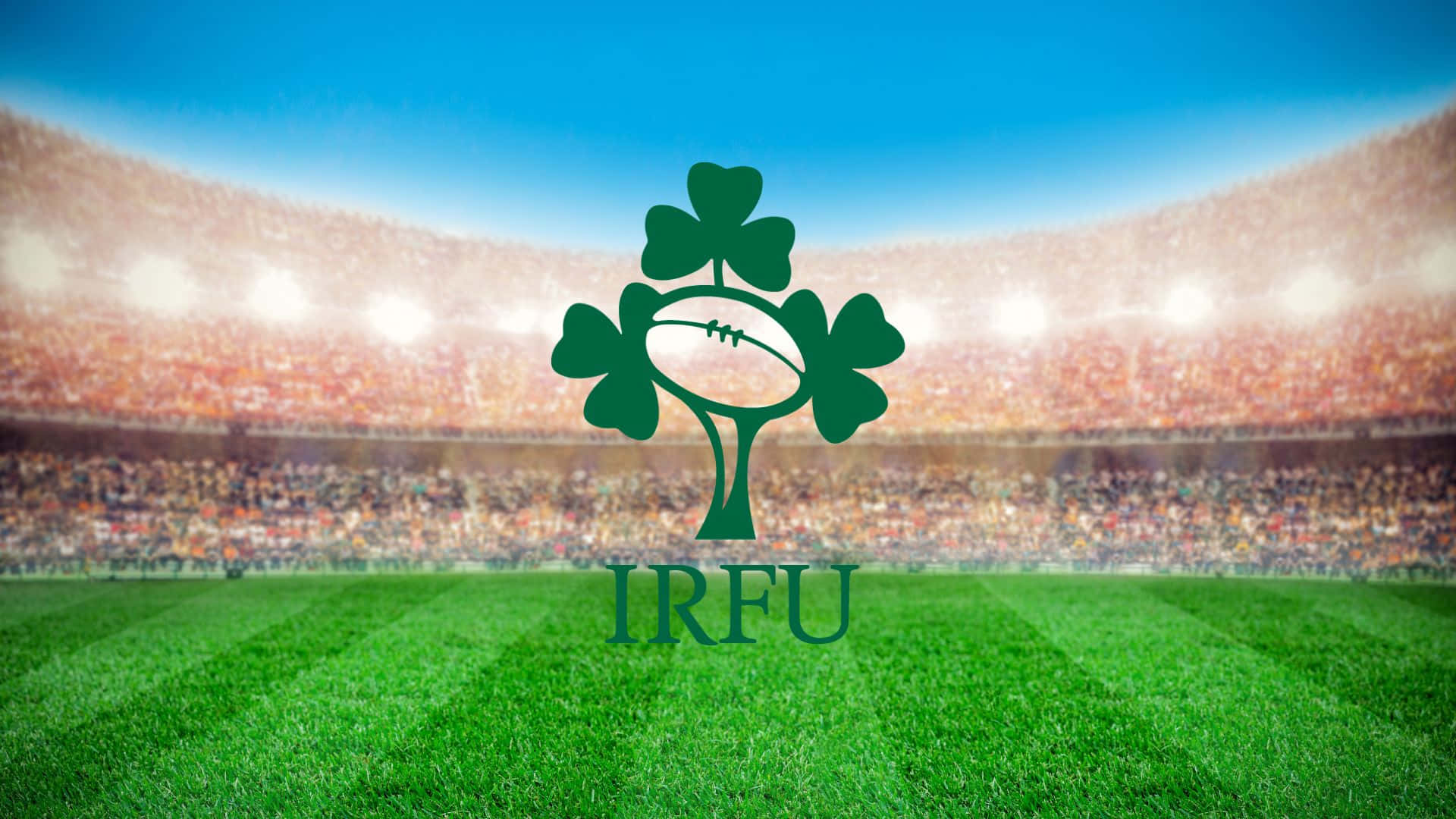 Ireland Rugby 桌布 壁紙 — 一個充滿活力的綠色三葉草標誌，中心有一個欖球，矗立在茂密的草地上，前方是座無虛席的體育場，天空湛藍清澈。
