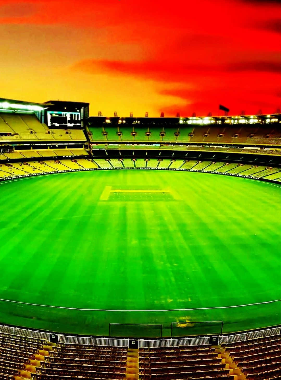 Cricket Ground 桌布 壁紙 — 一個充滿活力的綠色板球場，在火紅和橙色的日落天空下，球場周圍空無一人。