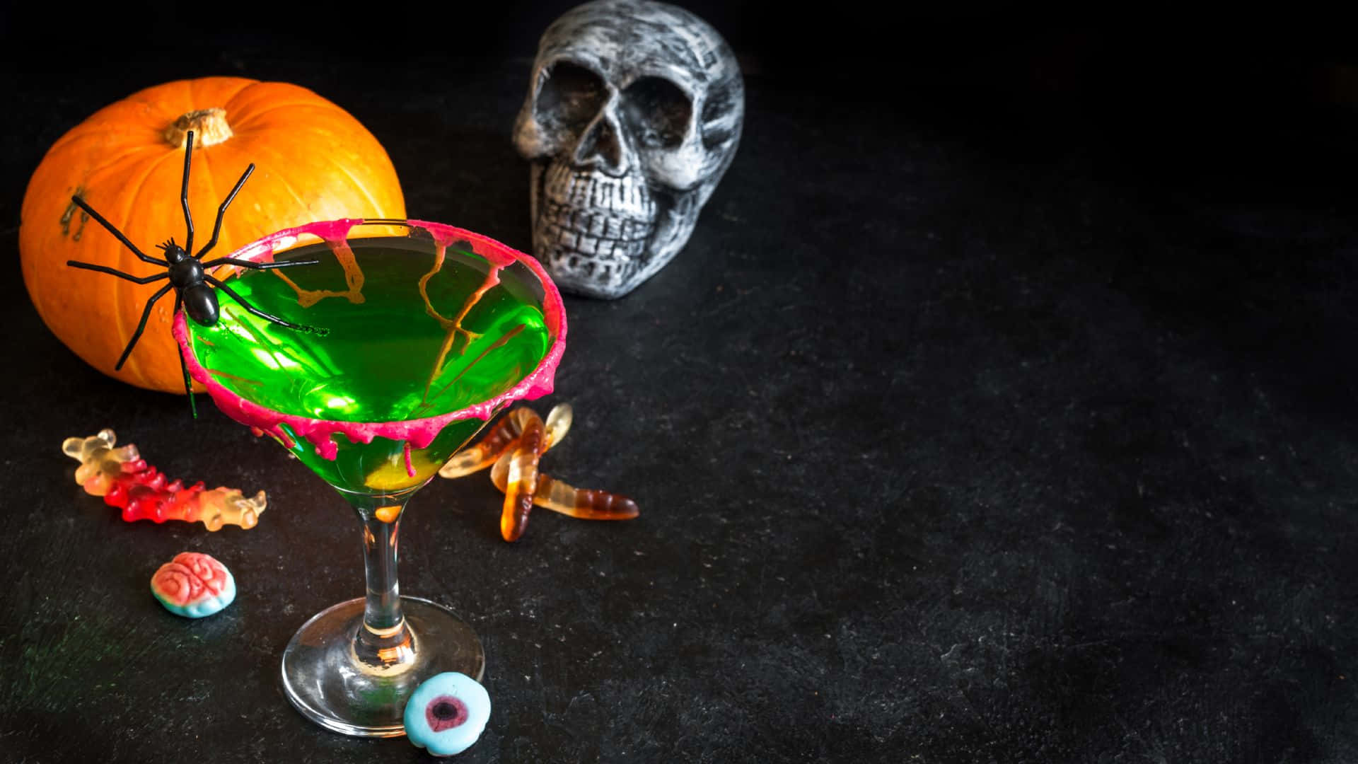 Spooky Cocktails 桌布 壁紙 — 一個充滿活力的綠色萬聖節雞尾酒，放在馬提尼杯中，周圍有南瓜、蜘蛛、骷髏頭、軟糖蟲和眼球，置於深色表面上。