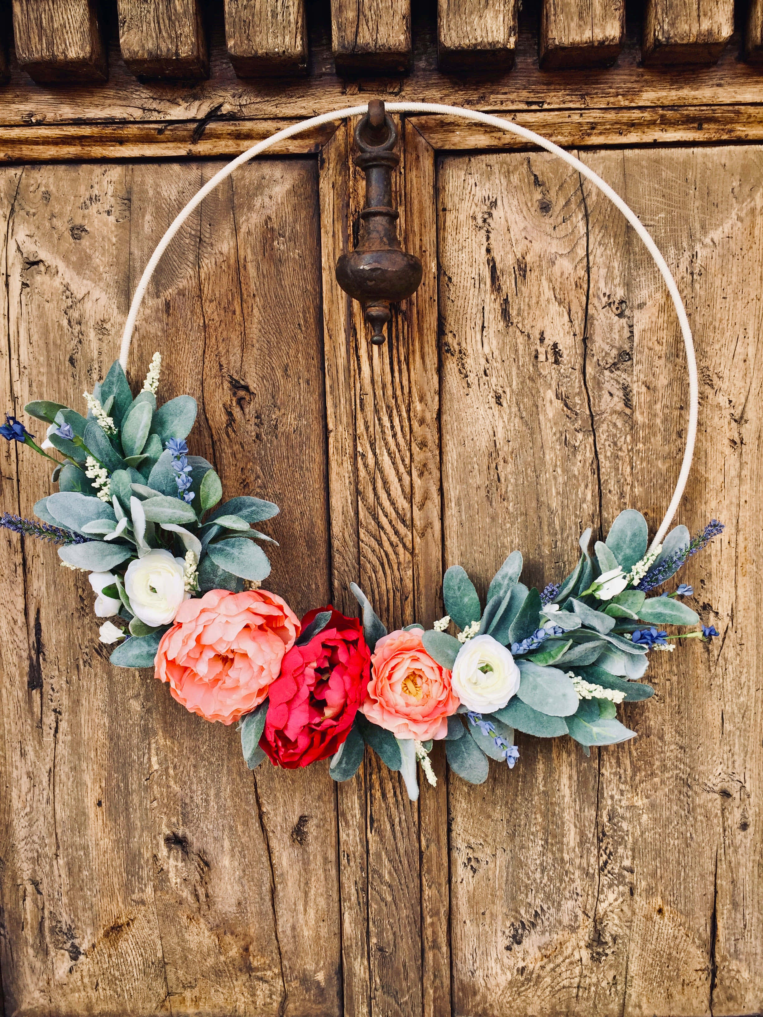Spring Wreaths 桌布 壁紙 — 一個充滿活力的花環，帶有珊瑚紅和蜜桃色的花朵，掛在一個帶有質樸金屬門環的風化木門上。