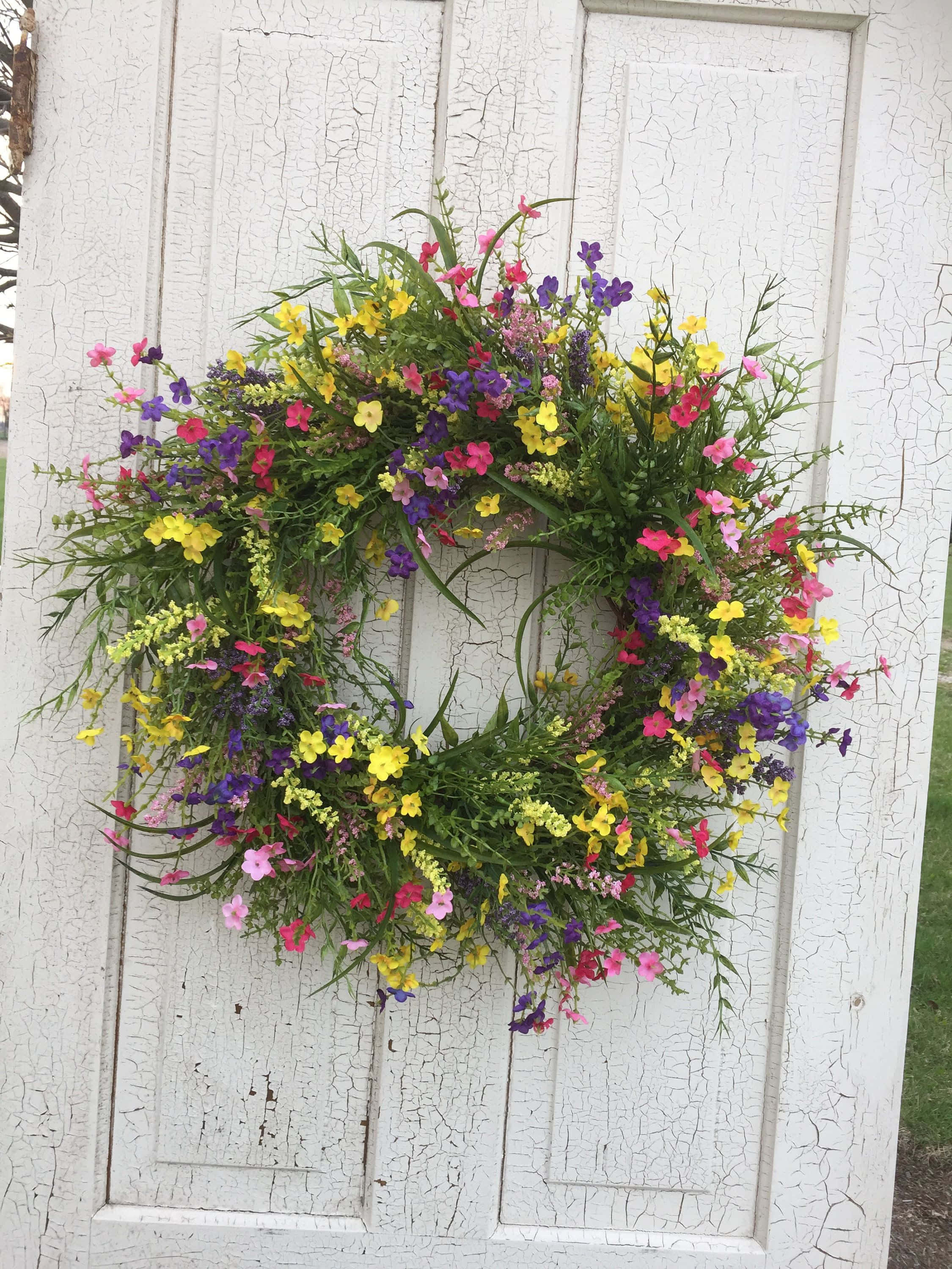 Spring Wreaths 桌布 壁紙 — 一個充滿活力的花環，綻放著粉紅色、黃色和紫色的花朵，掛在風化的白色木門上。