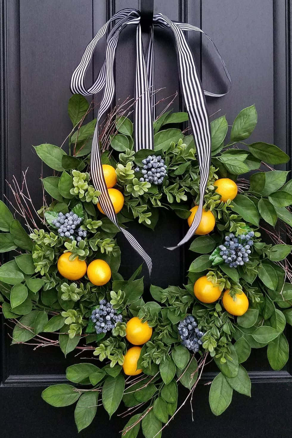 Spring Wreaths 桌布 壁紙 — 一個充滿活力的花環，飾有黃色柑橘類水果和藍色漿果，掛在深色木門上，用黑白條紋絲帶繫著。