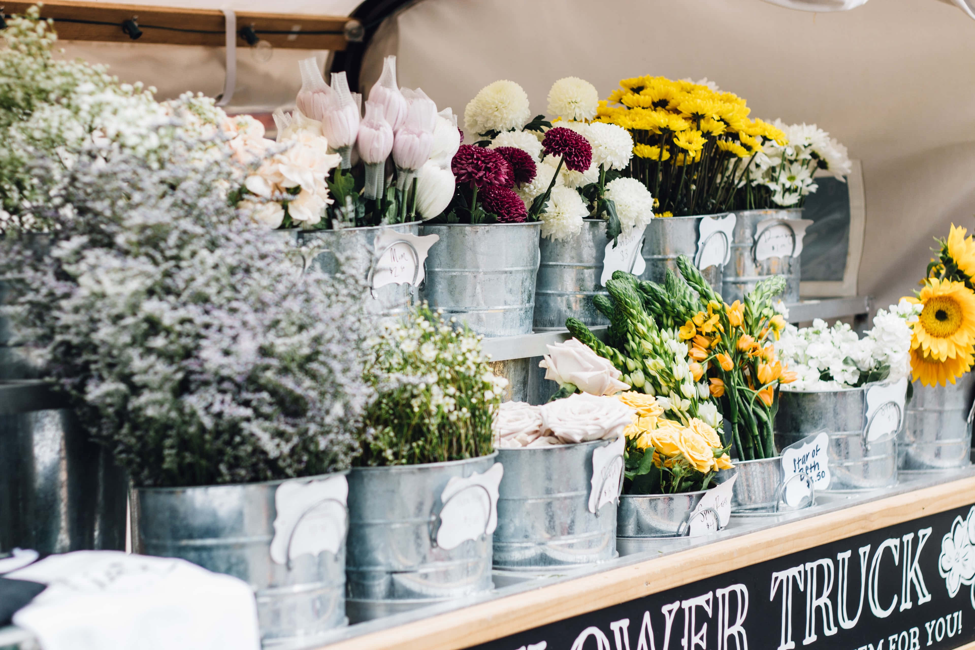 Flower Shop 桌布 壁紙 — 一個充滿活力的花車展示，鍍鋅桶中裝有粉紅色鬱金香、黃色雛菊和白色花朵，置於遮陽篷下。