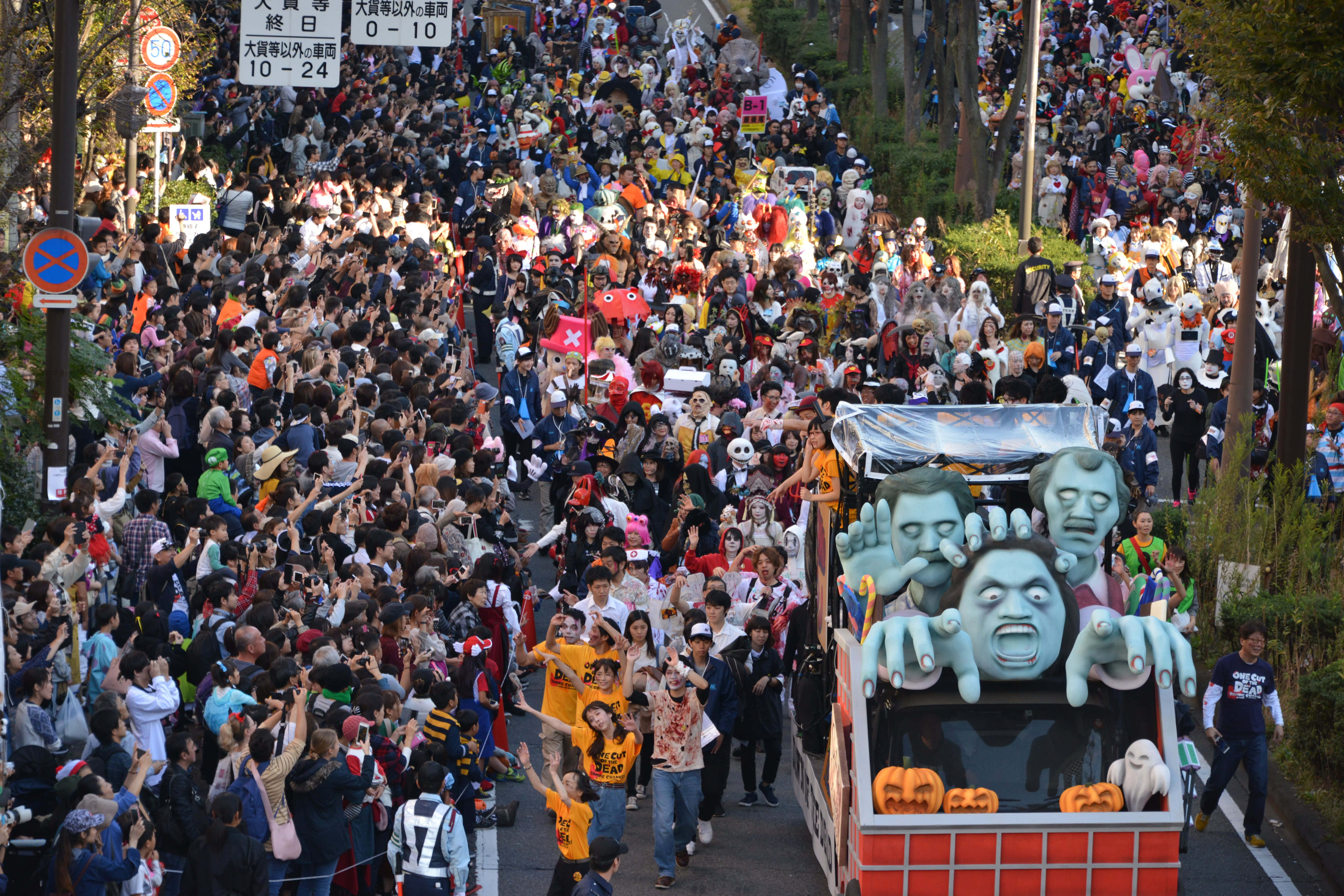Halloween Parades 桌布 壁紙 — 一個充滿活力的萬聖節巡遊，擠滿了樹木成蔭的街道，盛裝打扮的人群歡呼著，一輛以殭屍為主題、裝飾著發光南瓜的花車緩緩駛過。