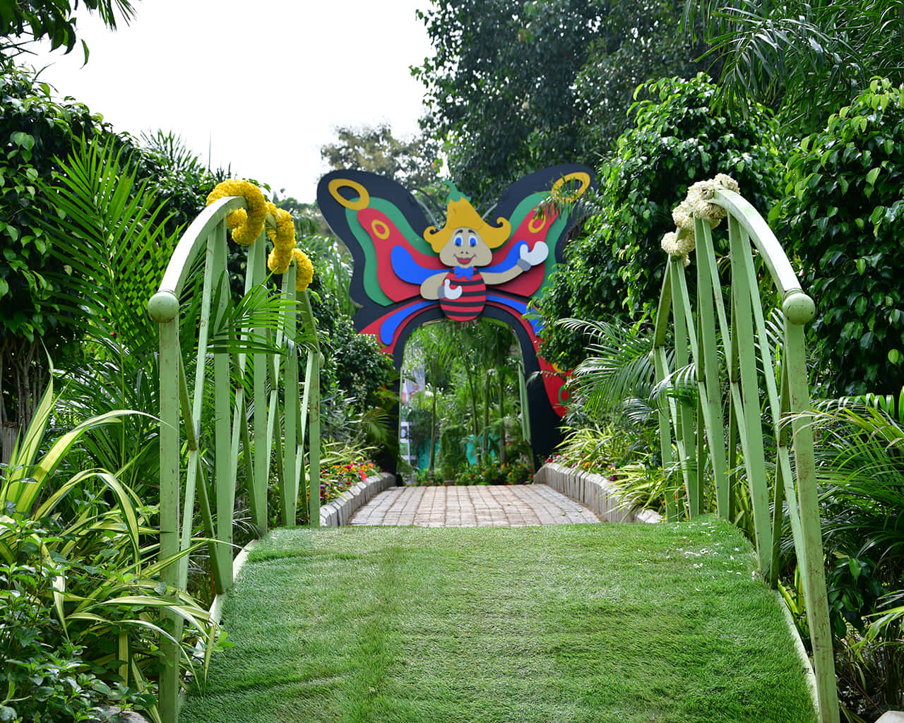 Butterfly Park 桌布 壁紙 — 一個充滿活力的蝴蝶形拱門，帶有卡通角色，歡迎訪客穿過茂密的綠色花園小徑。