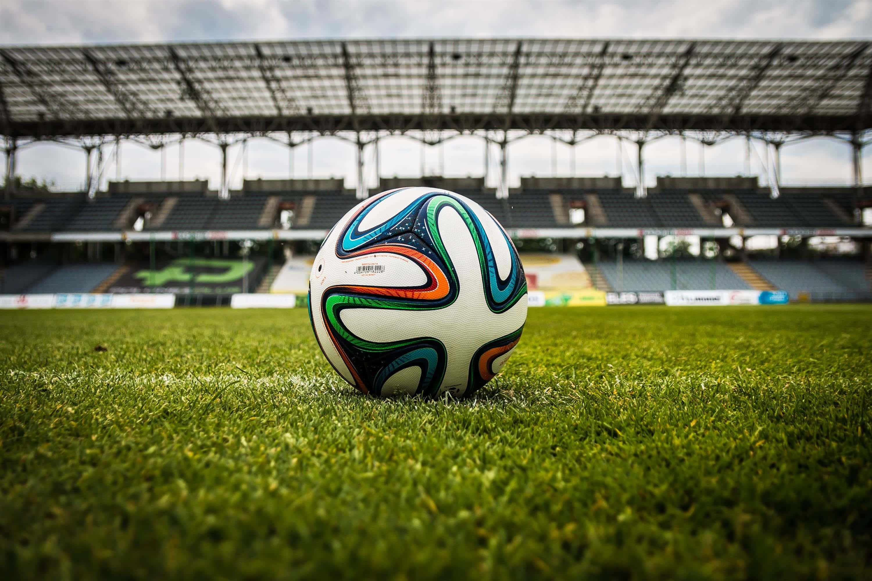 Soccer Stadium 桌布 壁紙 — 一個充滿活力的足球放在空蕩蕩的體育館內茂密的綠草上，天空多雲。