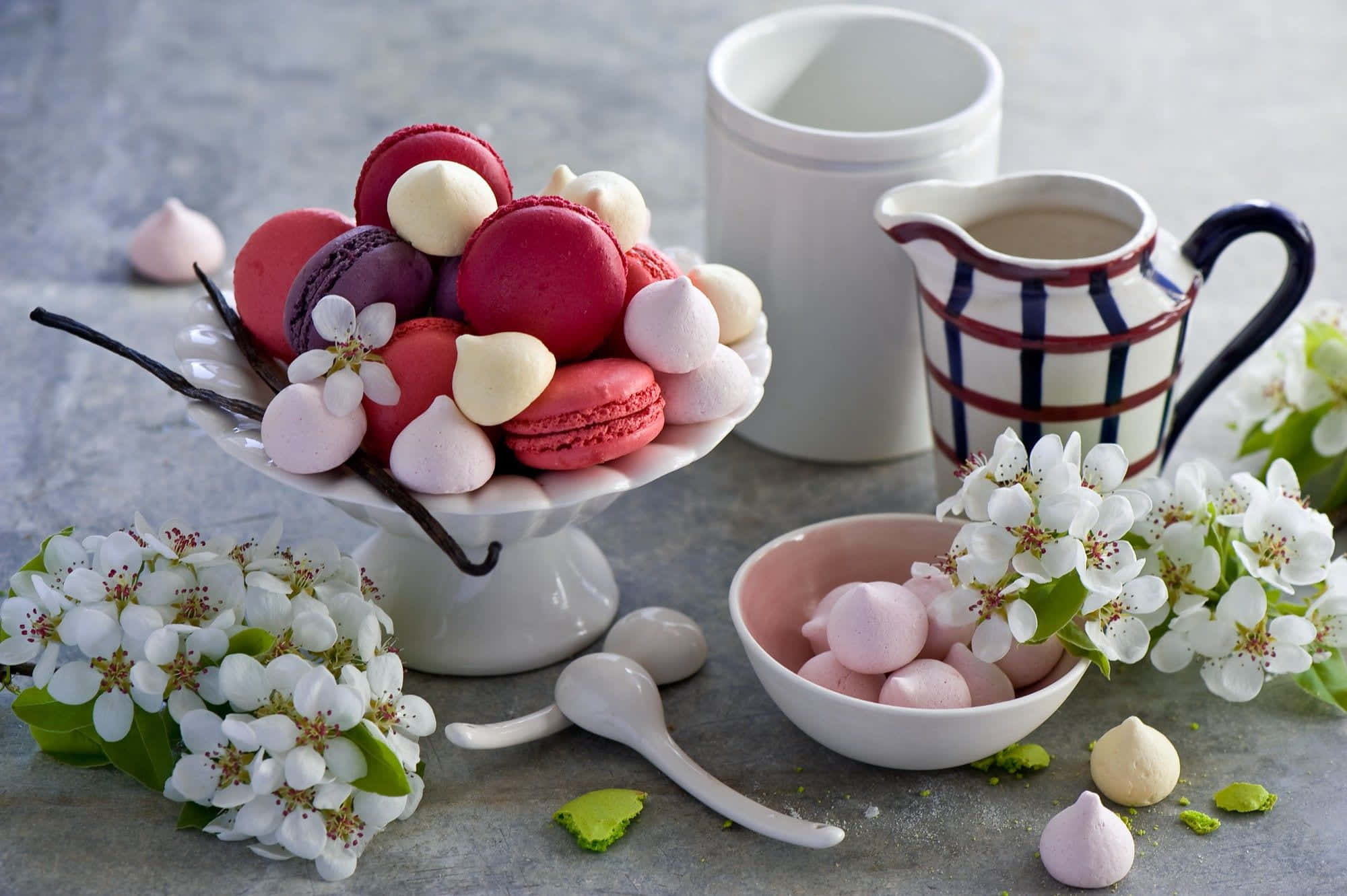 Spring Desserts 桌布 壁紙 — 一個充滿活力的靜物，白色碗中盛放著色彩繽紛的馬卡龍和蛋白霜，旁邊是盛開的白花和一個條紋水壺。