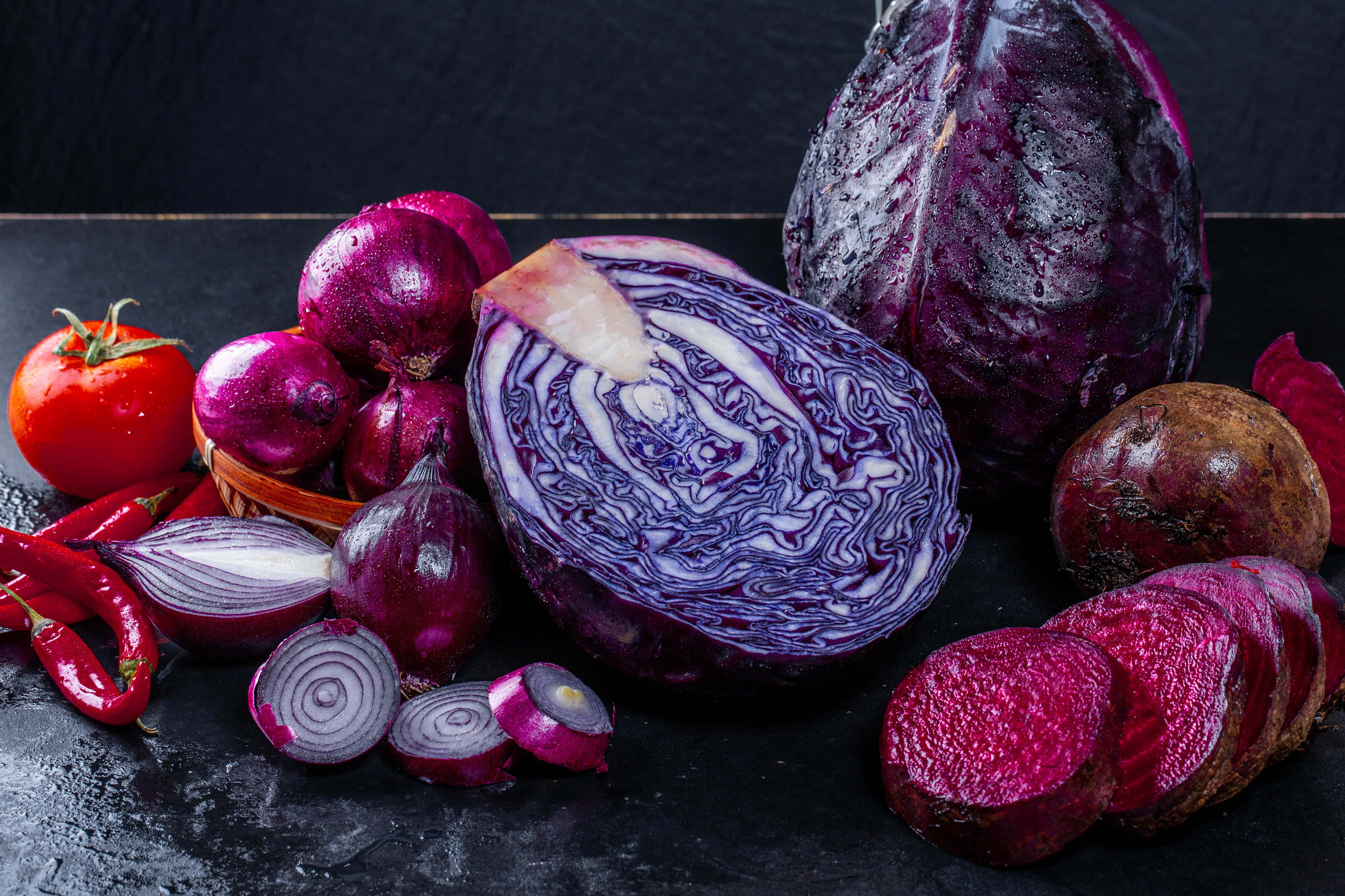 Purple Onions 桌布 壁紙 — 一個充滿活力的靜物畫，展示了切片紅椰菜、紅菜頭、車厘茄和紅洋蔥，排列在深色紋理表面上。