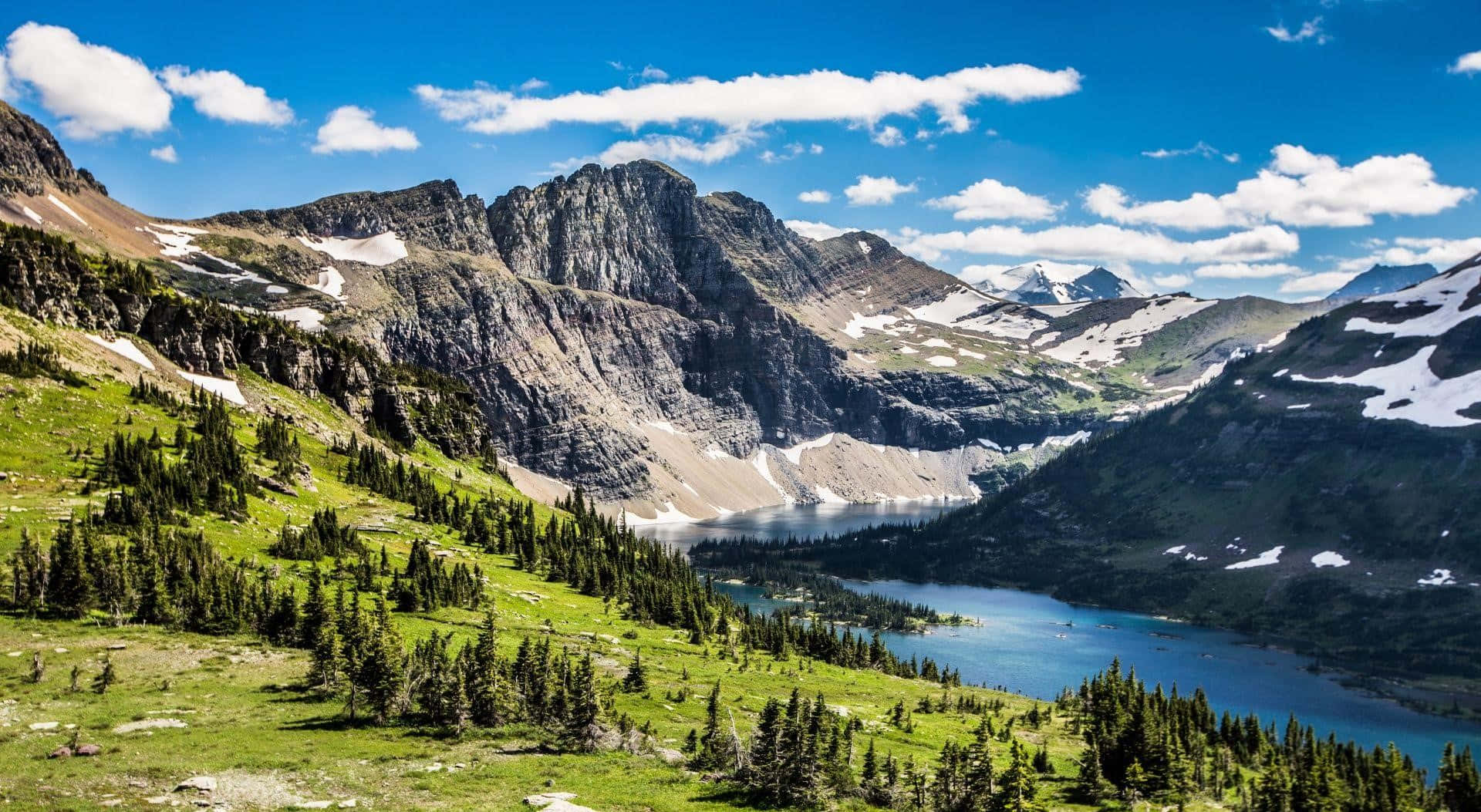 Montana 桌布 壁紙 — 一個充滿活力的高山湖泊，坐落在崎嶇的積雪山脈之間，上方是明亮的藍天和蓬鬆的白雲。