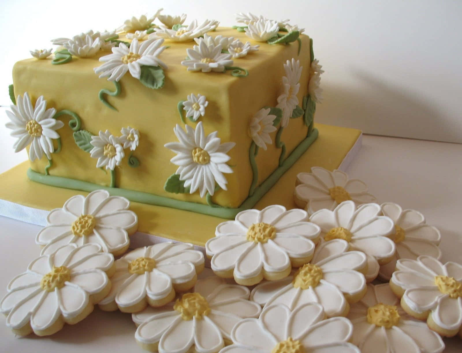 Spring Desserts 桌布 壁紙 — 一個充滿活力的黃色長方形蛋糕，裝飾著白色雛菊和綠色藤蔓，旁邊放著一堆相配的雛菊形狀餅乾。