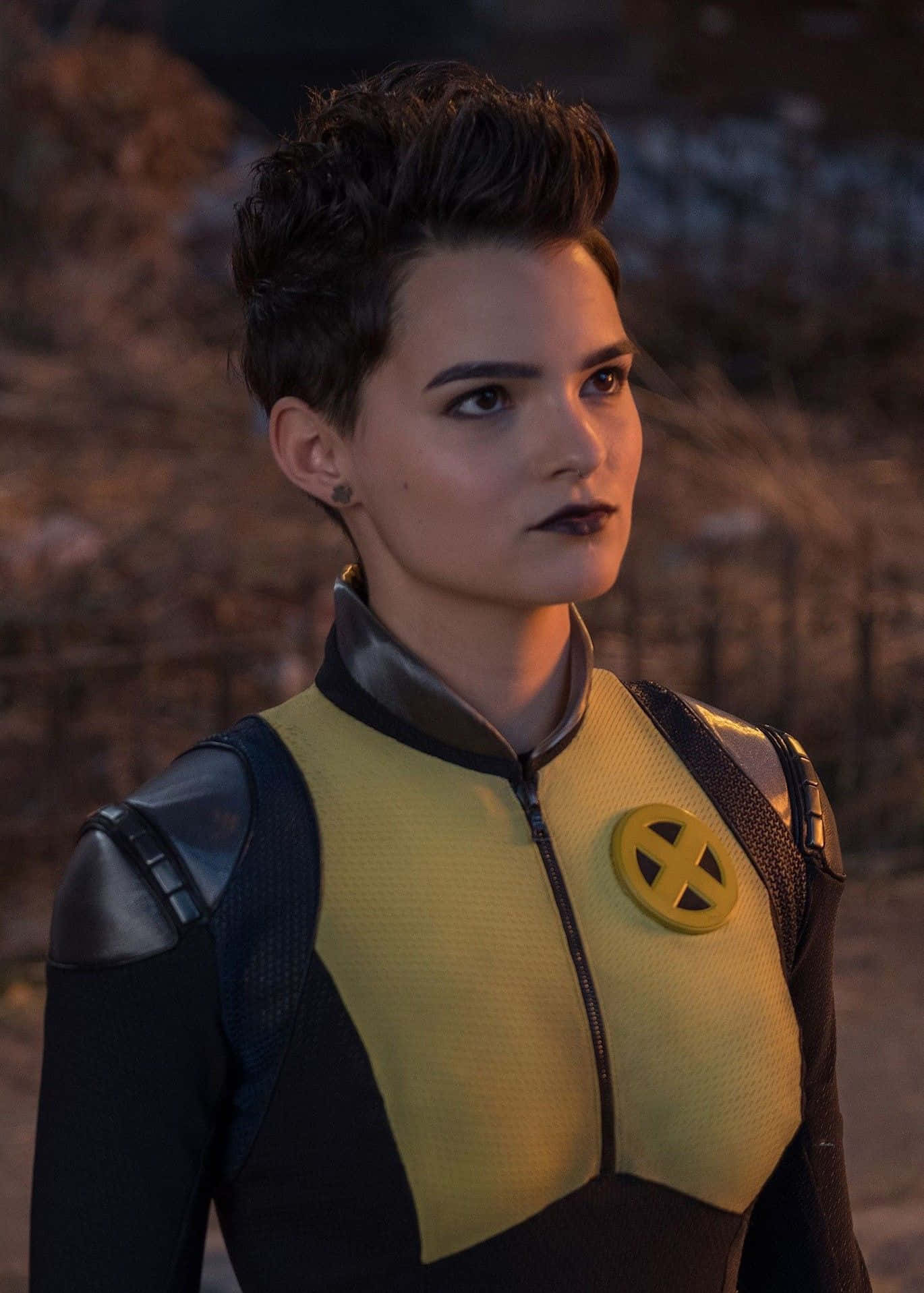 Negasonic Teenage Warhead 桌布 壁紙 — 一個兇猛的變種人穿著黃黑相間的X-Men戰衣，在昏暗的戶外環境中神情嚴肅地站立。