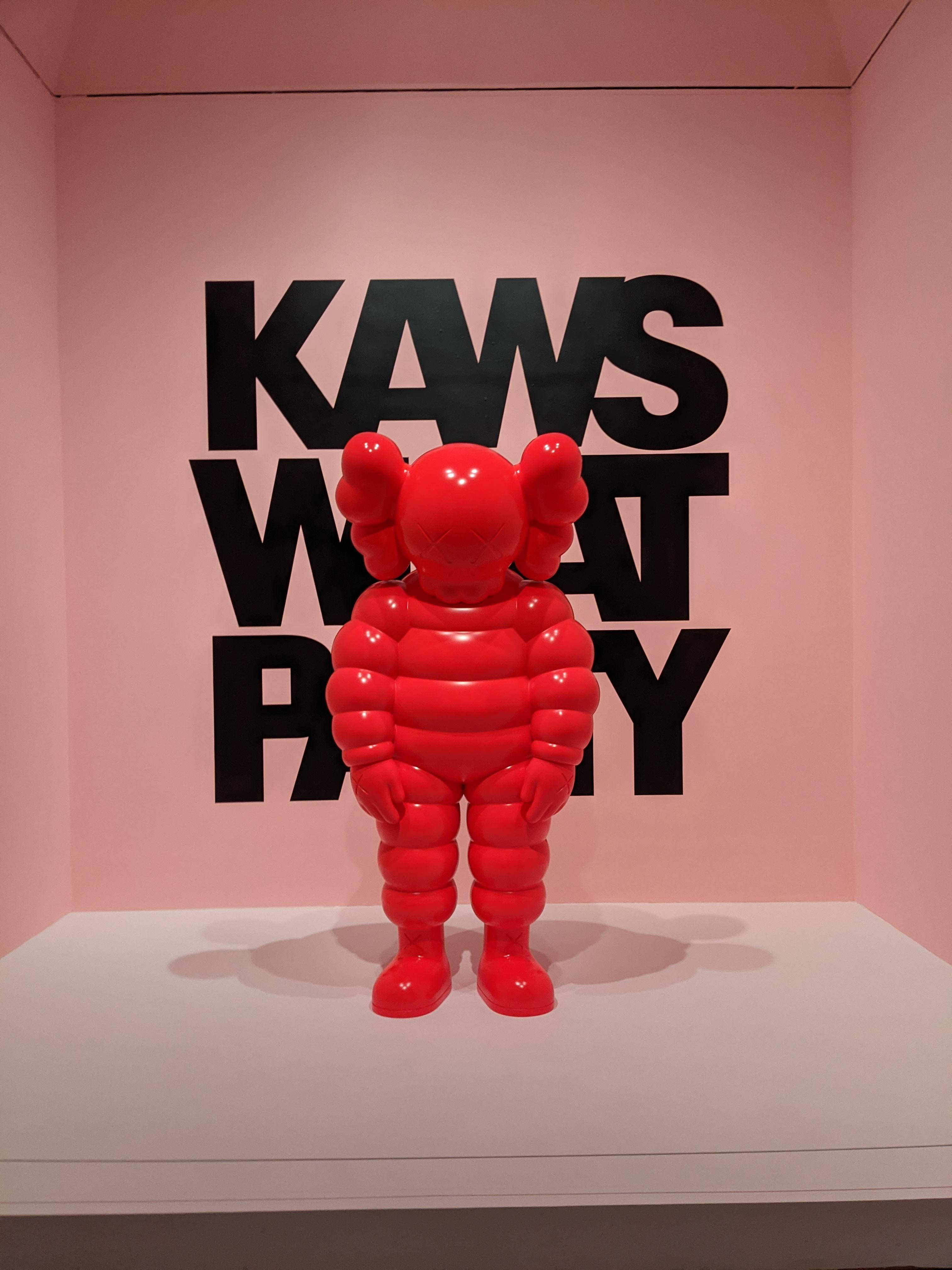 KAWS What Party 壁紙 — 一個光澤的紅色KAWS Companion雕塑立在白色底座上，背景是粉紅色的牆壁，上面有粗體黑色'KAWS WHAT PARTY'文字。