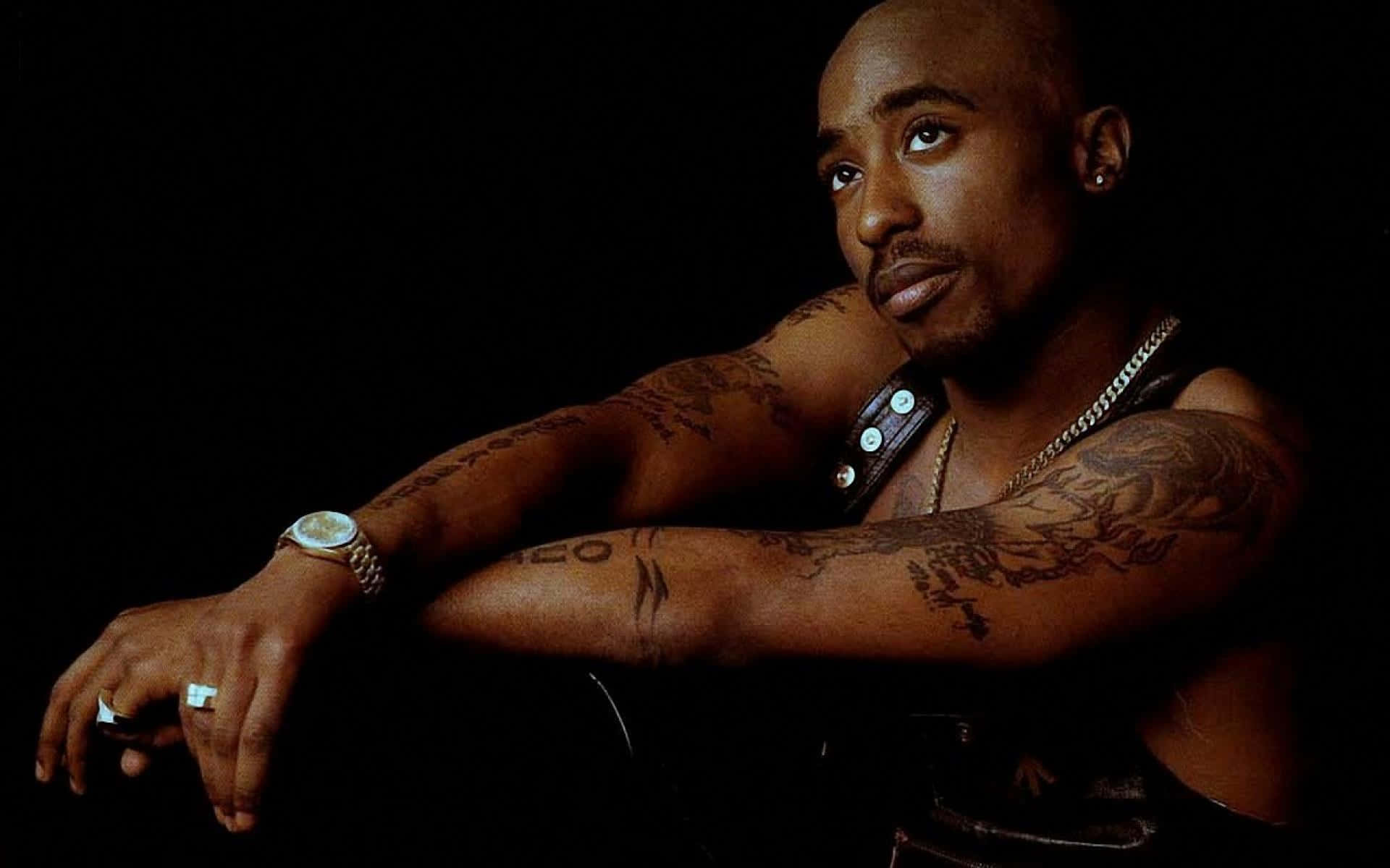Tupac 桌布 壁紙 — 一個光頭、手臂有紋身的男人，在黑色背景前用手托住下巴，戴著手錶和鏈條。