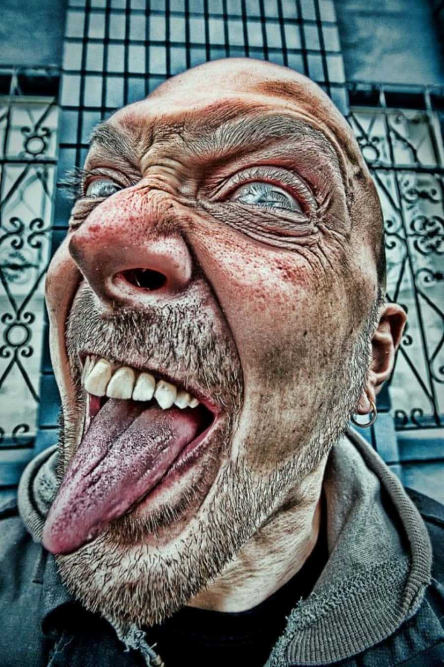 Funny Faces 桌布 壁紙 — 一個光頭男人做出怪誕誇張的鬼臉，伸出舌頭，背景是藍色金屬圍欄。