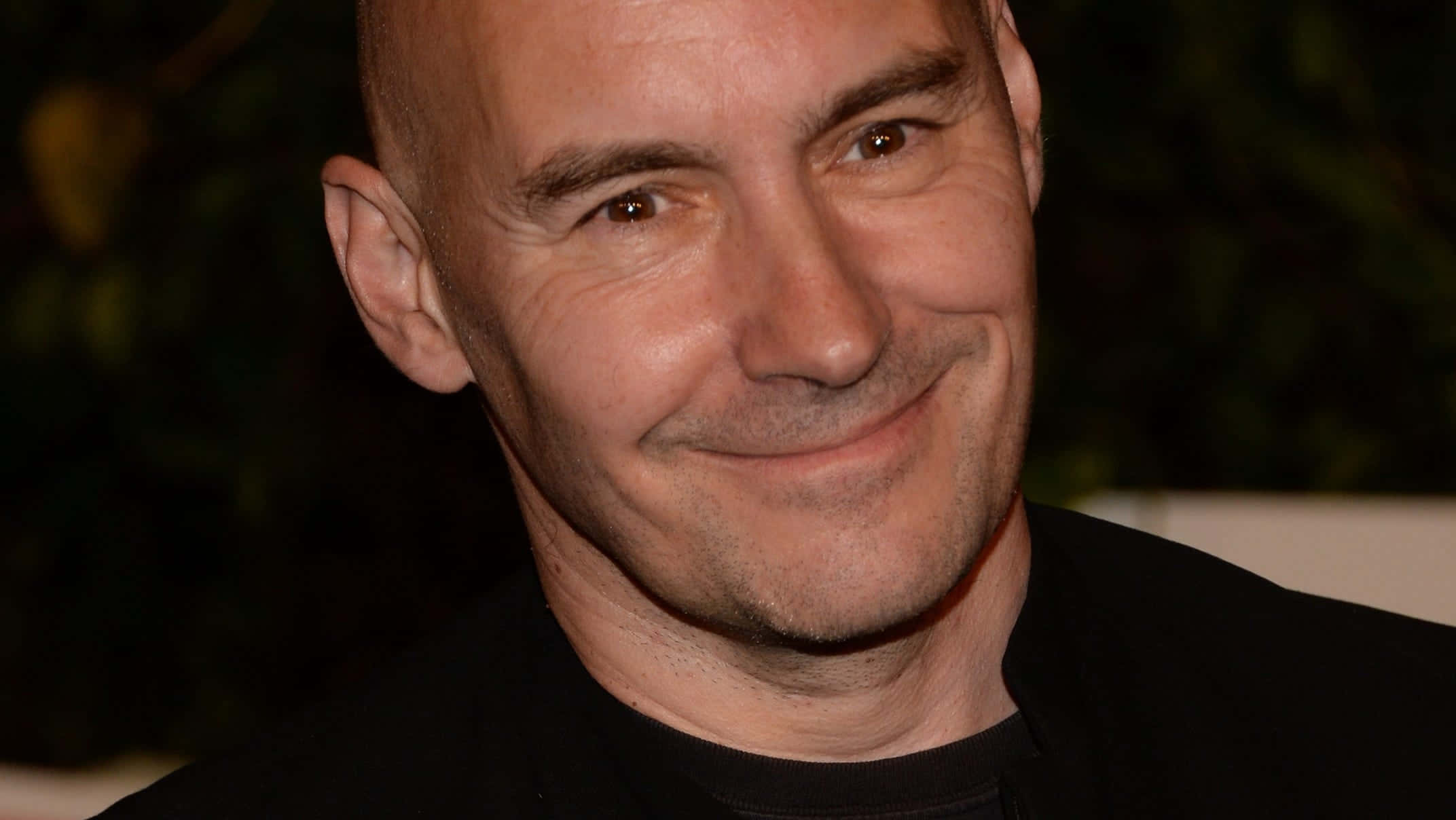 Grant Morrison 桌布 壁紙 — 一個光頭男人嘅近距離肖像，帶住溫暖嘅笑容，著住黑色恤衫，背景係暗色模糊嘅。