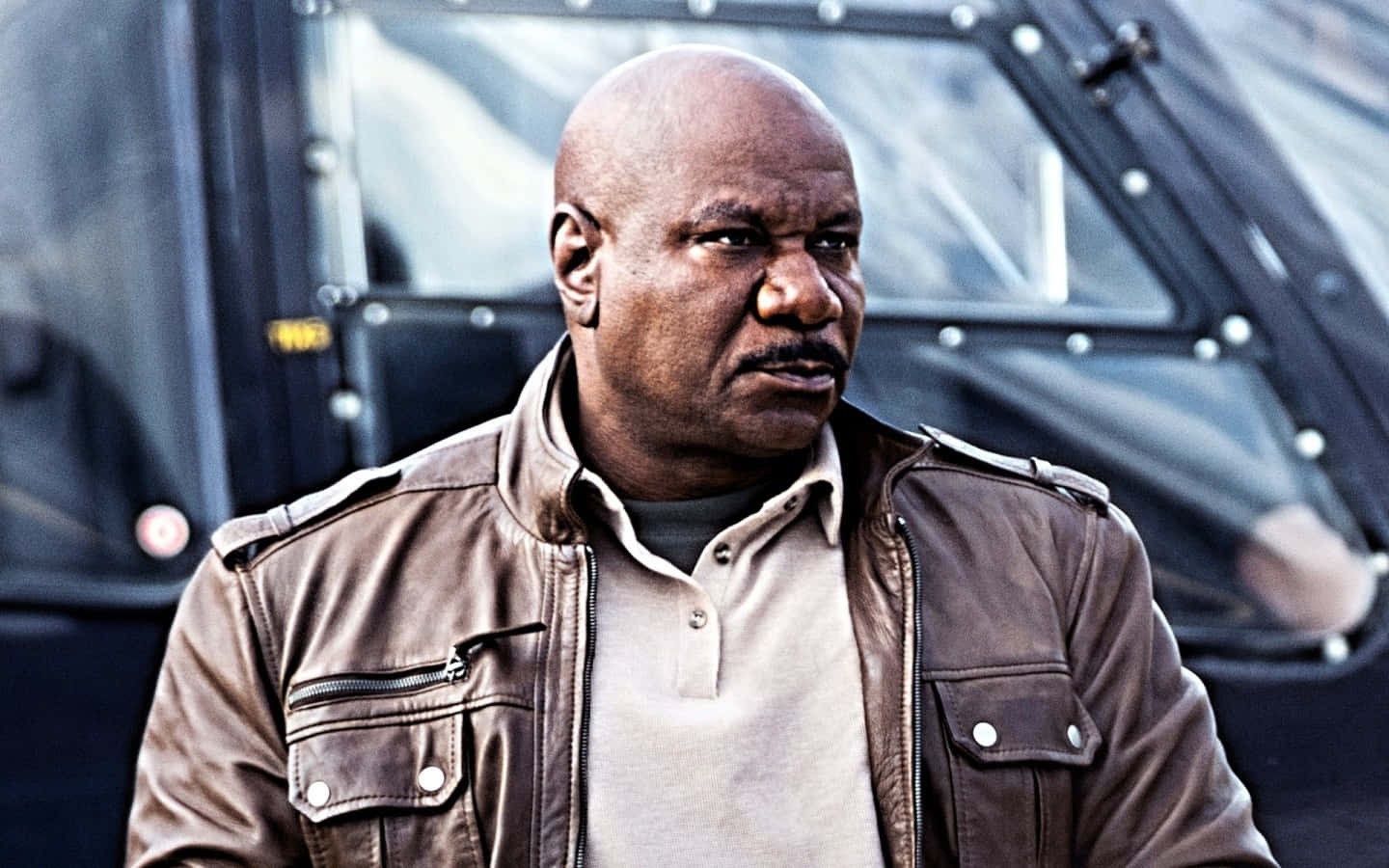 Ving Rhames 桌布 壁紙 — 一個光頭男人穿著棕色皮褸，神情嚴肅地站在一輛黑色裝甲車前。
