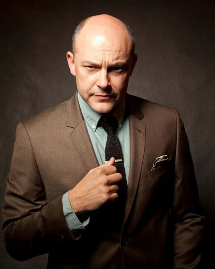 Rob Corddry 桌布 壁紙 — 一個光頭男人穿著棕色西裝，在黑暗的攝影棚背景下，戲劇性的燈光中調整領帶。