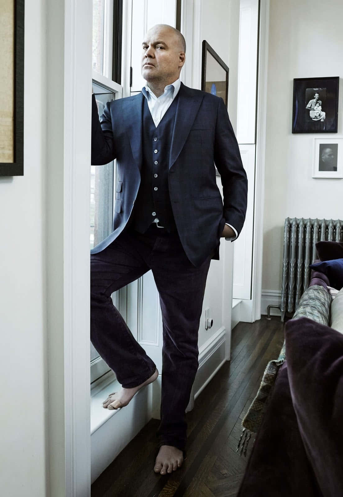 Vincent D'onofrio 桌布 壁紙 — 一個光頭男人穿著深色西裝，赤腳站在窗台上，在一個舒適且充滿藝術品的房間裡若有所思地向外凝望。