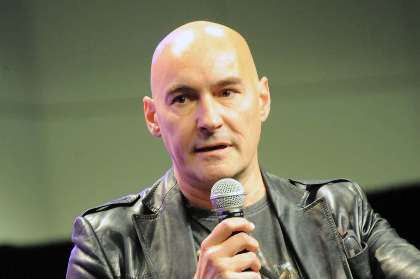 Grant Morrison 桌布 壁紙 — 一個光頭男人穿著黑色皮褸，專注地對著咪高峰說話。