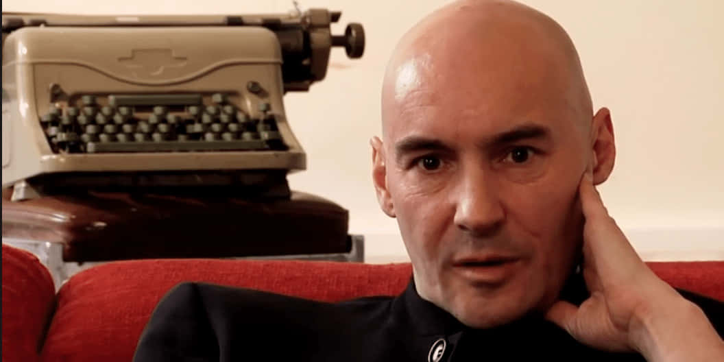 Grant Morrison 桌布 壁紙 — 一個光頭男人穿著黑色襯衫，手托下巴坐在紅色梳化上，身後有一部復古米色打字機。