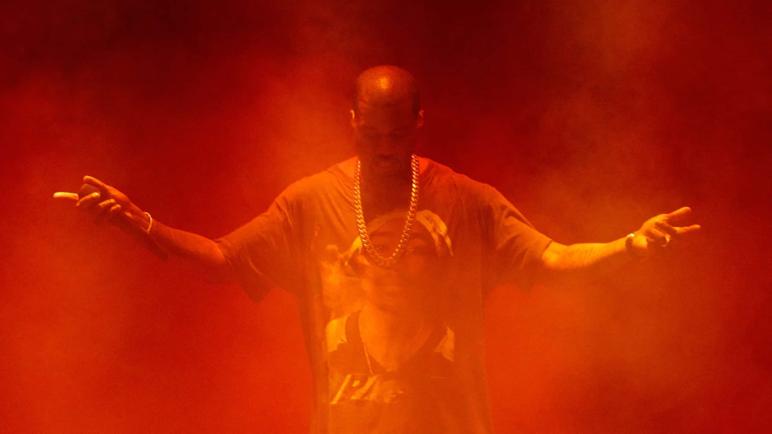 Kanye 桌布 壁紙 — 一個光頭男子穿著圖案T恤和鏈條，在煙霧瀰漫的紅色舞台環境中張開雙臂站立。