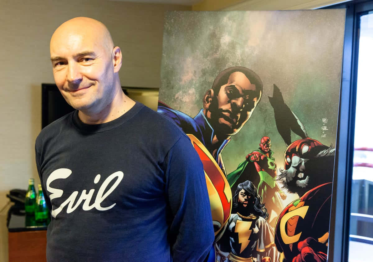 Grant Morrison 桌布 壁紙 — 一個光頭男子穿著藍色 'Evil' T恤，在房間裡一張大型漫畫風格的超級英雄海報旁微笑。