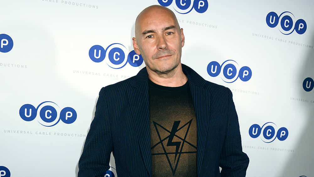 Grant Morrison 桌布 壁紙 — 一個光頭男子身穿深色細條紋西裝外套和圖案T恤，自信地站在帶有品牌標誌的UCP背景前。