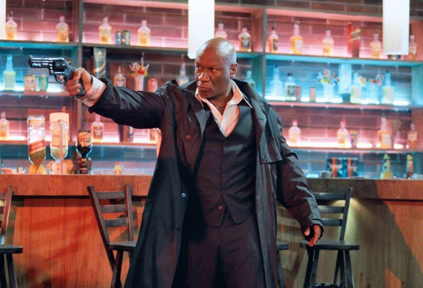Ving Rhames 桌布 壁紙 — 一個光頭男子身穿黑色乾濕褸，在燈光昏暗、身後有發光酒架的酒吧中瞄準左輪手槍。