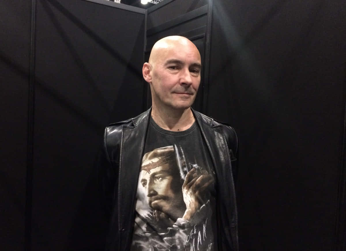 Grant Morrison 桌布 壁紙 — 一個光頭男子身穿黑色皮褸和圖案T恤，站在柔和燈光的黑暗背景下。