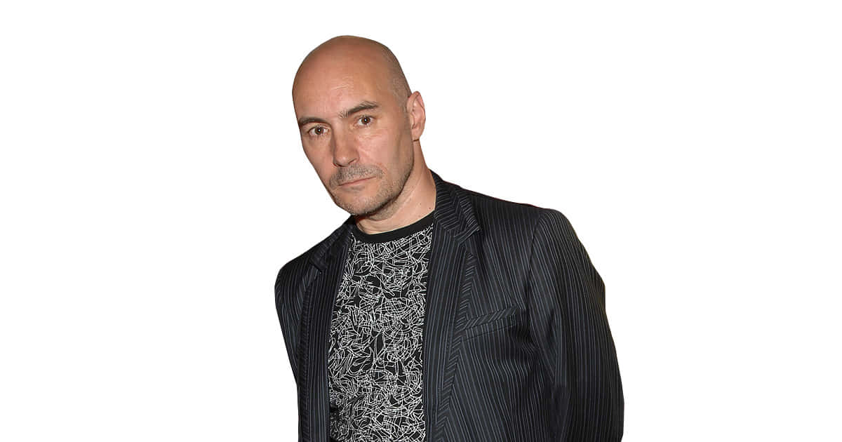 Grant Morrison 桌布 壁紙 — 一個光頭留山羊鬍的男人，在純白背景前穿著一件條紋西裝外套，內搭圖案襯衫。