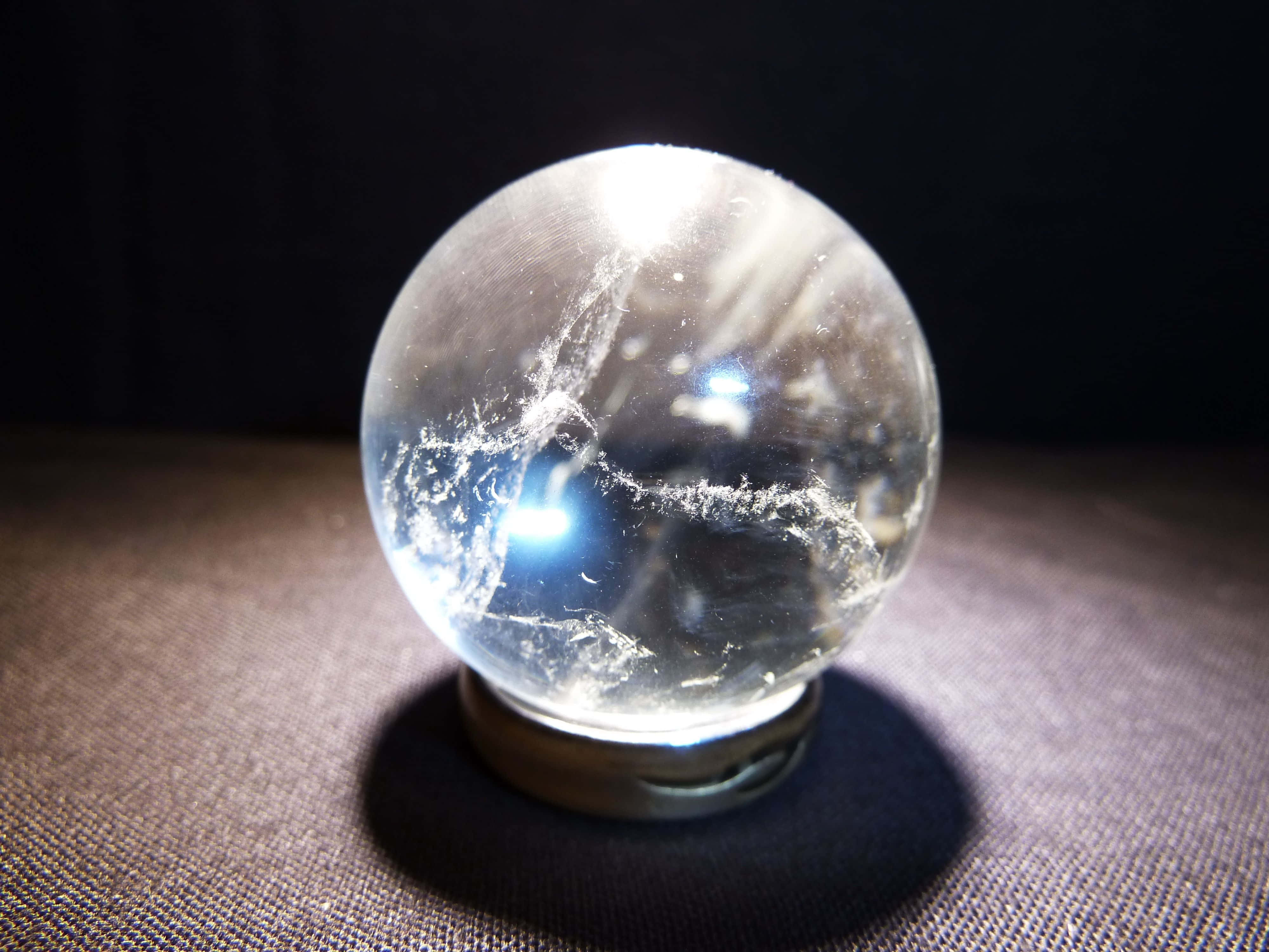 Crystal Ball 桌布 壁紙 — 一個內部有裂紋的透明水晶球，放置在金屬底座上，背景昏暗，由明亮光源照亮。