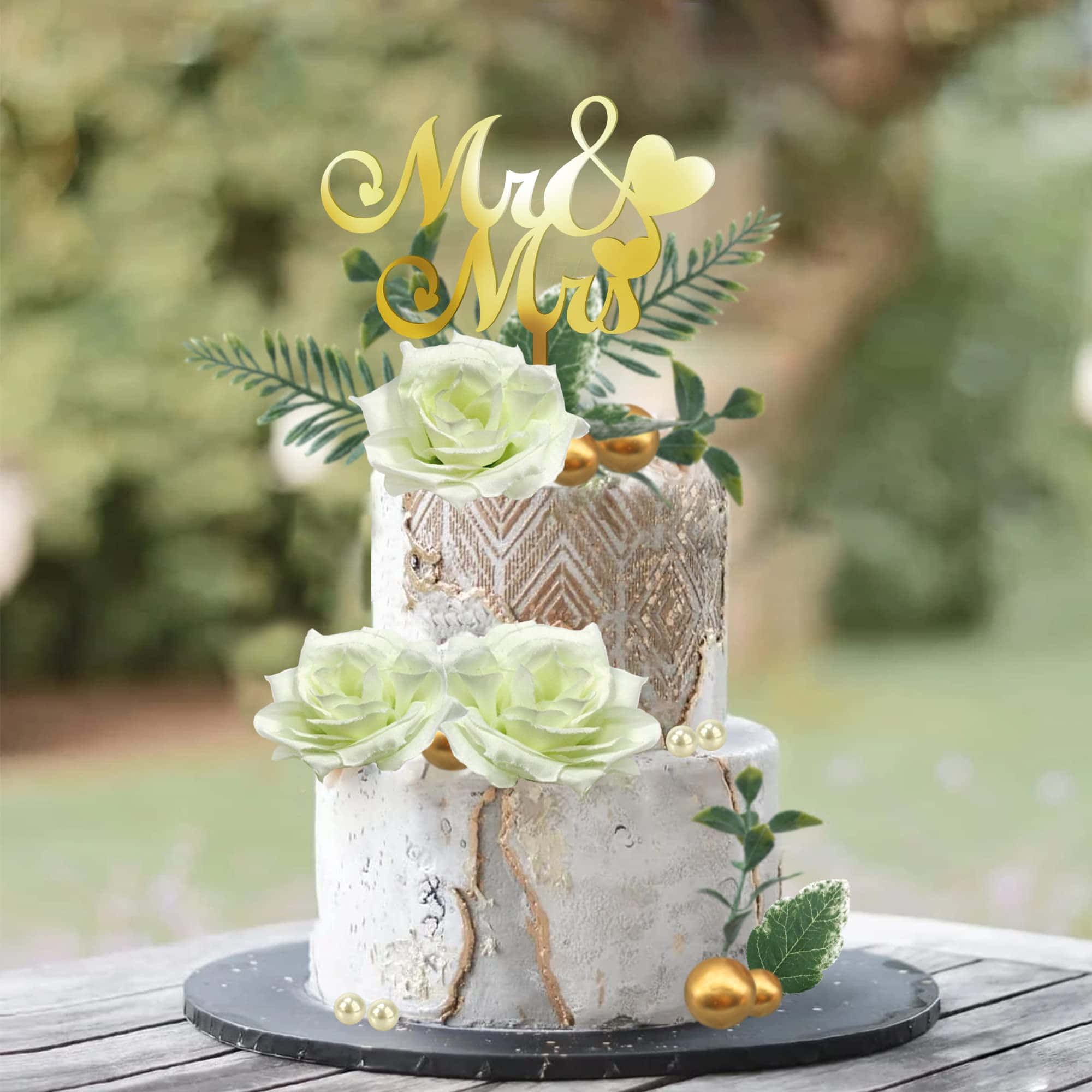 Floral Cake 桌布 壁紙 — 一個兩層高的婚禮蛋糕，上面有白玫瑰、金色裝飾和 'Mr & Mrs' 裝飾，放在戶外的木桌上。
