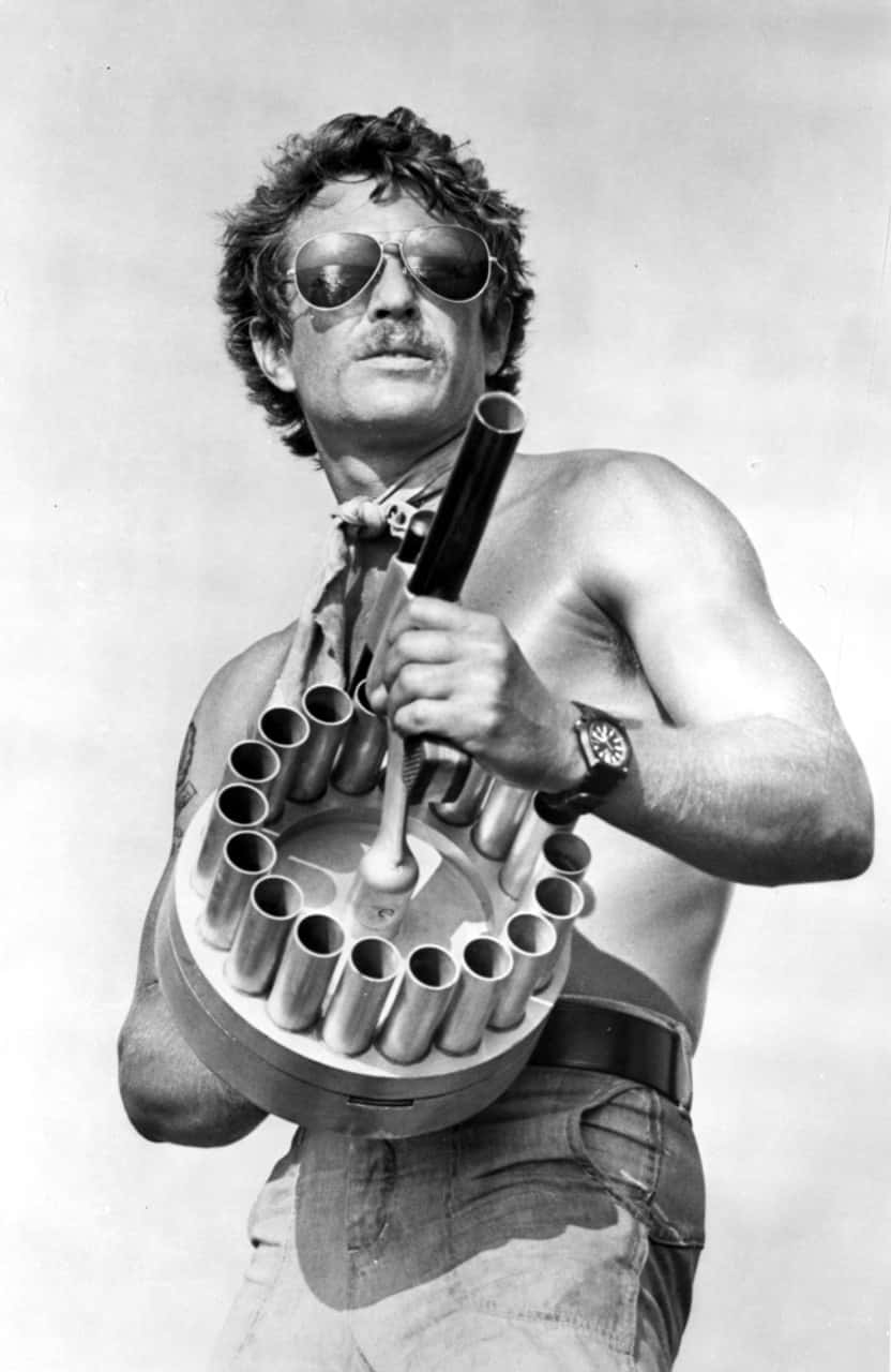 Tom Berenger 桌布 壁紙 — 一個冇著衫嘅男人戴住太陽眼鏡同鬍鬚，喺普通天空背景下揸住一把大型多管武器。