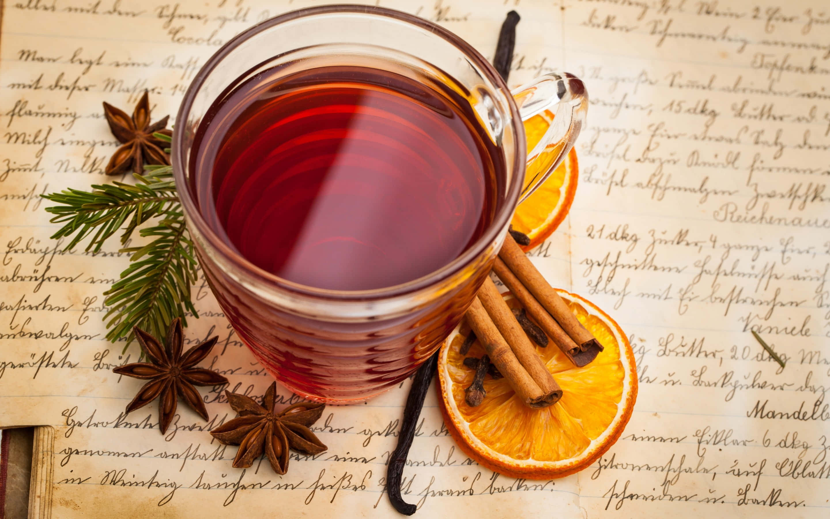Mulled Wine 桌布 壁紙 — 一個冒煙的玻璃杯，裝著加了肉桂棒、橙片和八角香料茶，放在手寫食譜紙上。