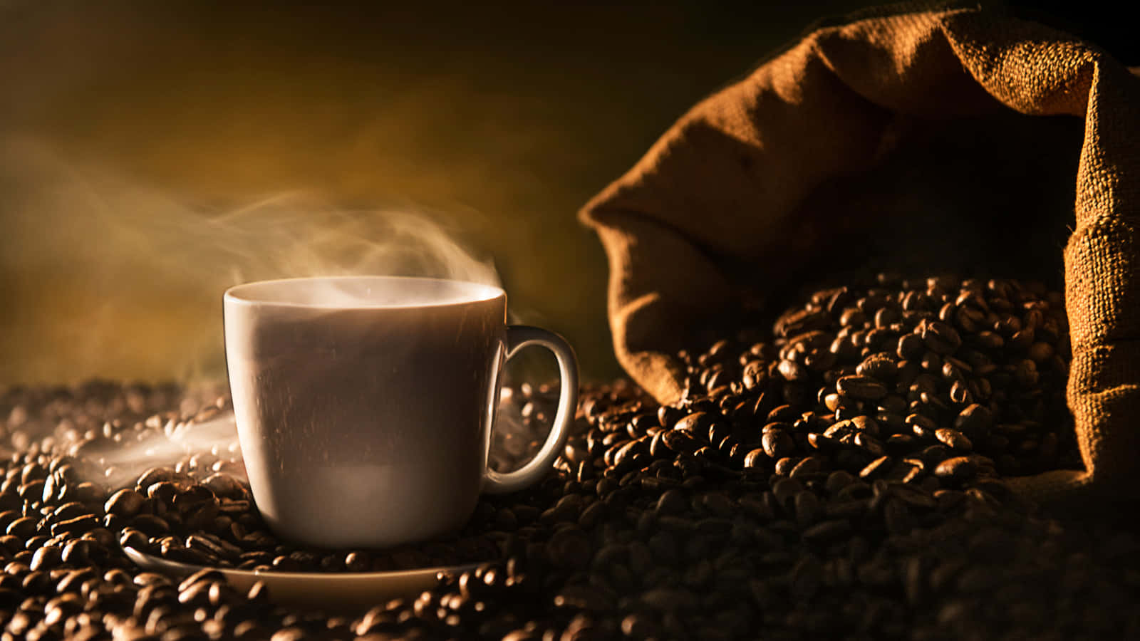 Brown Coffee 桌布 壁紙 — 一個冒煙的白色咖啡杯放在深烘焙咖啡豆中，附近有一個麻布袋，在溫暖的金色燈光下。
