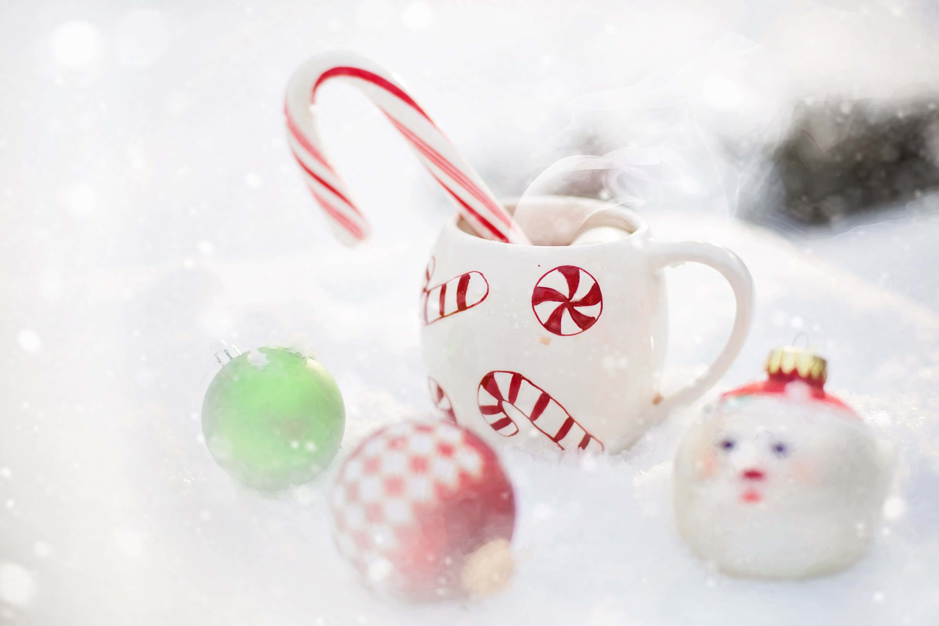 Hot Chocolate 桌布 壁紙 — 一個冒著蒸氣、帶有拐杖糖圖案的杯子放在雪中，旁邊有節日裝飾品和一根拐杖糖。