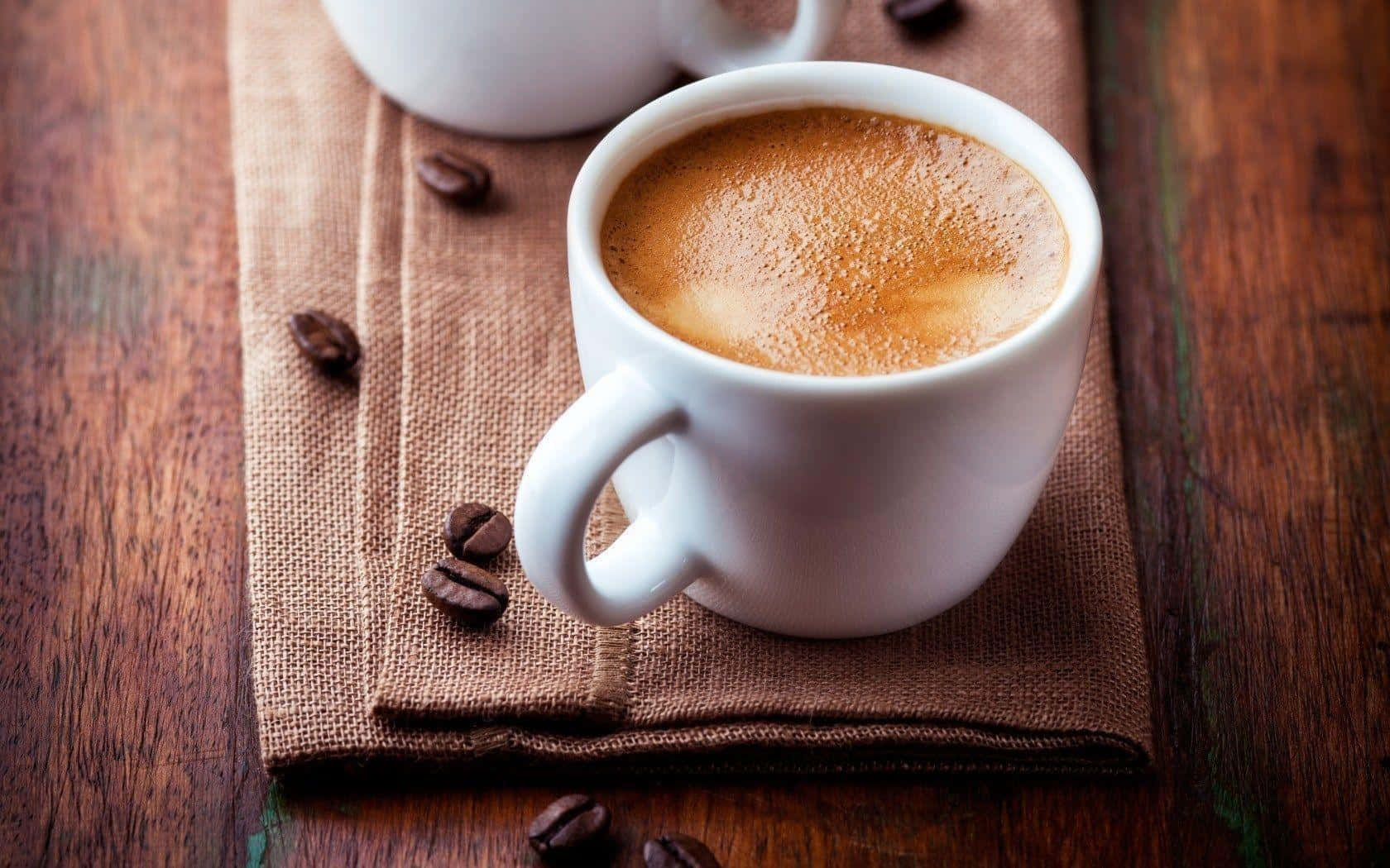 Coffee Cup 桌布 壁紙 — 一個冒著蒸氣的白咖啡杯，帶有泡沫奶泡，放在棕色麻布餐巾上，周圍散落著咖啡豆，置於鄉村風木桌上。